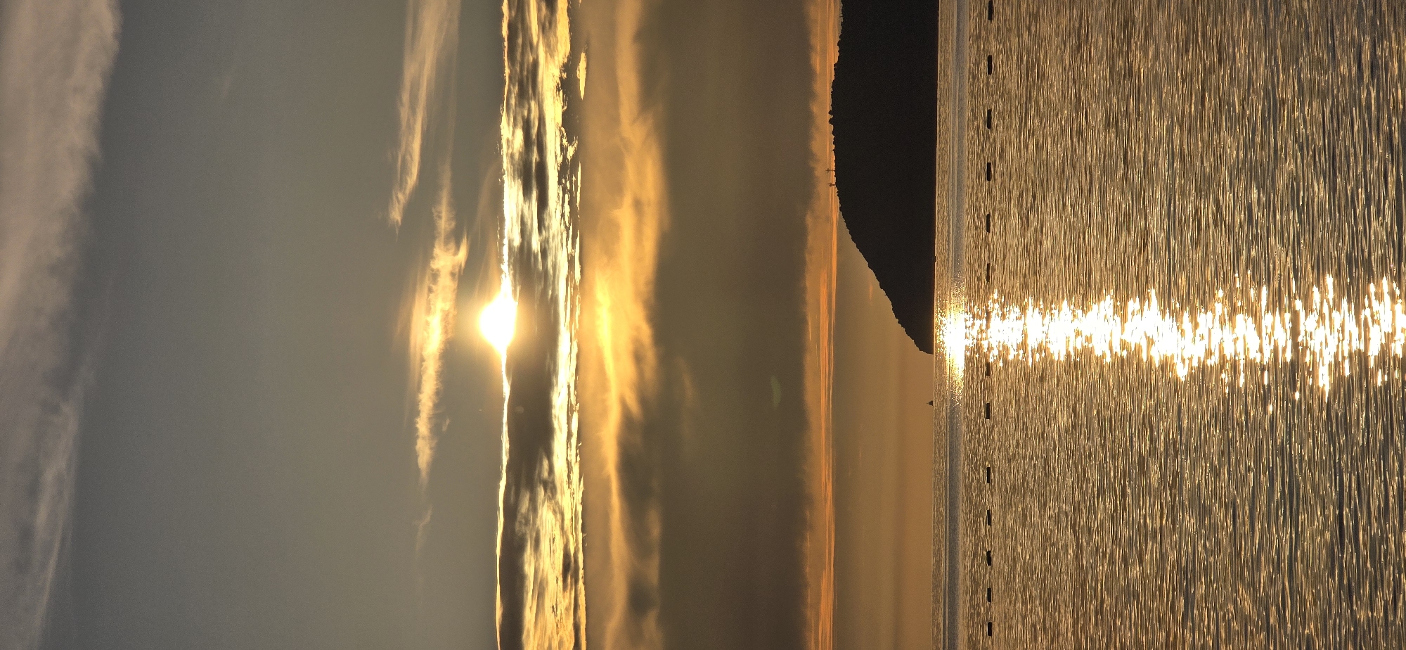 A vertical photograph taken in Bar, Montenegro, captures a tranquil maritime sunrise or sunset. The sun, a bright golden orb, is centered in the upper third of the frame, partially obscured by horizontal bands of clouds that are illuminated in vivid orange and yellow hues, contrasting with the darker grey clouds above. The lower two-thirds of the image depict the calm sea, reflecting the sun's radiance as a prominent vertical streak of shimmering golden light extending from the horizon to the foreground. A linear arrangement of approximately ten dark, cylindrical buoys or markers floats horizontally across the mid-ground water. On the right side of the horizon, a dark, silhouetted landmass, likely a hill or peninsula, rises from the water, displaying two faint vertical structures on its peak. A small, dark vessel is discernible on the water near the base of the landmass. The overall color palette is dominated by warm golden tones, with natural light and atmospheric conditions as the primary subjects. No human figures are present.