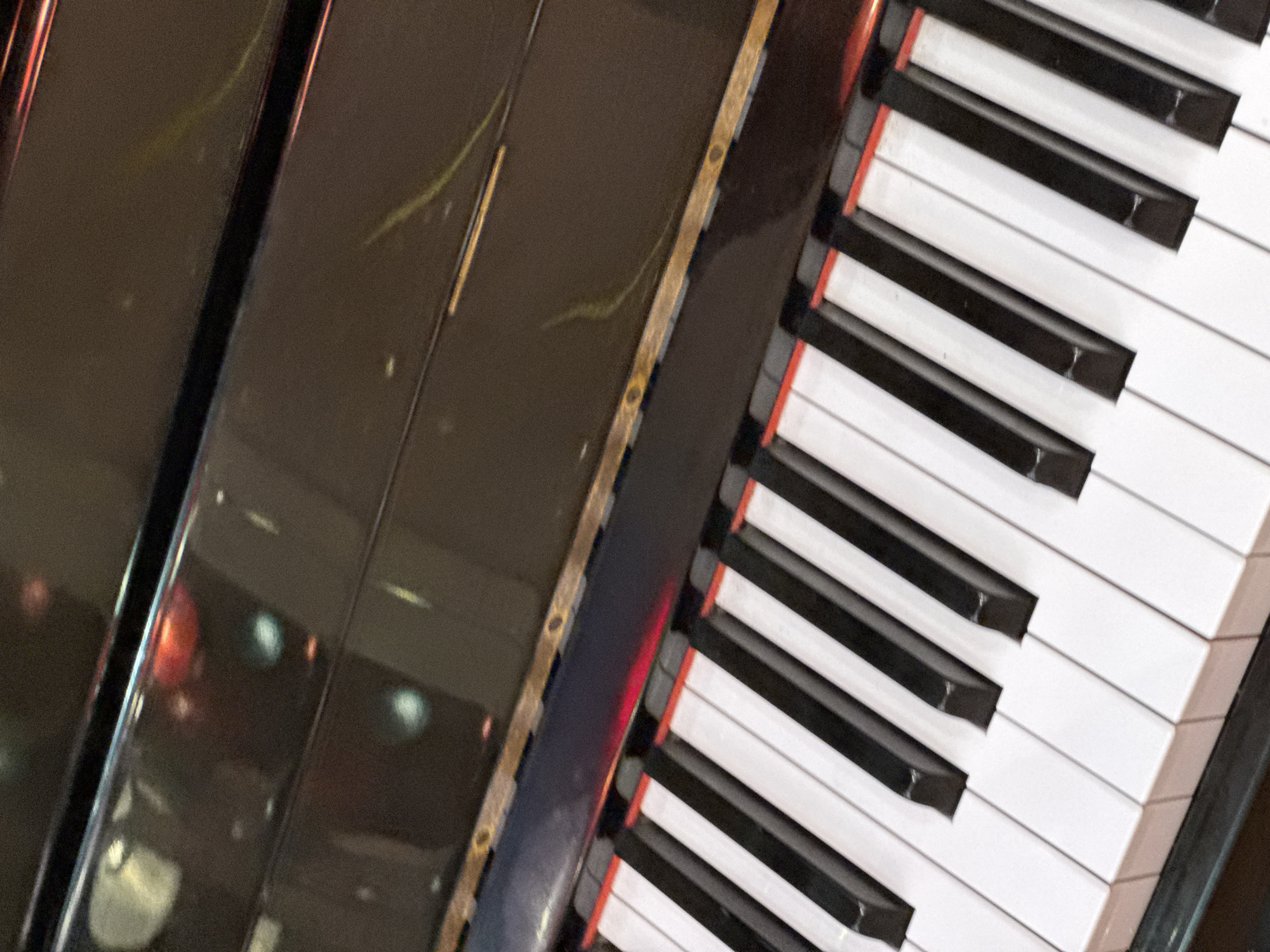The image is a close-up, angled shot of the keys of an upright piano. The keys are white and black, with red accents visible between the white keys in some areas. The piano has a glossy black finish, reflecting light and indistinct shapes, possibly from surrounding lighting or furniture. The background of the piano's surface appears dark and reflective. The scene is devoid of any people or specific environmental context that would allow for identification of its location in Nha Trang Ward, Vietnam.