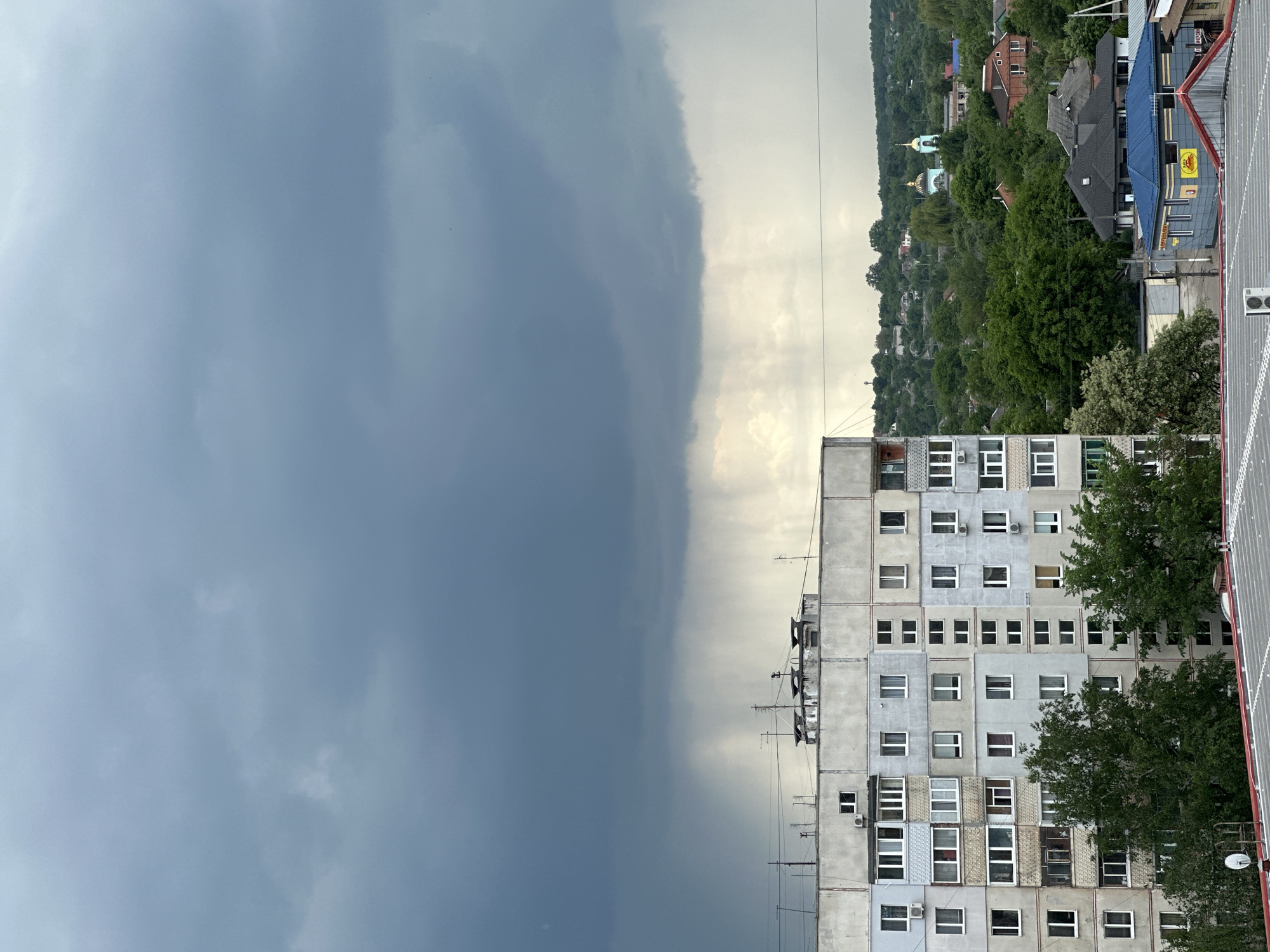 The image captures an urban scene under a dramatically divided sky. The left side is dominated by a menacing, dark gray storm cloud, while the right side is brighter, showing a lighter sky with some visible sun rays. The boundary between these two weather systems is sharply defined.
In the foreground, a tall, Soviet-era apartment building stands, its gray concrete facade punctuated by numerous windows. Some windows have air conditioning units installed. Two trees, displaying dense green foliage, partially obscure sections of the building.
Beyond the apartment building, a distant, undulating landscape is visible, covered with trees and interspersed with houses. The rooftops of these houses are varied in color, and a distinctive green dome of a church can be seen nestled among the foliage.
The image is taken from an elevated perspective, possibly from another building, offering a wide view of the city and the contrasting weather conditions. No people are visible in the image.