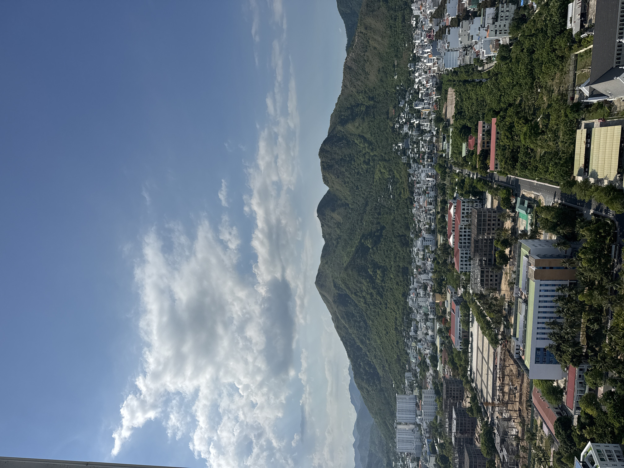 This is a high-angle, wide shot of a city nestled against a backdrop of lush green mountains under a bright blue sky with scattered white clouds. The foreground features a construction site with the skeletal frames of several buildings, indicating ongoing development. Adjacent to the construction, several completed multi-story buildings with white facades and red-tiled roofs are visible. A paved road winds through the scene, lined with trees. The middle ground shows a densely populated urban area with numerous white buildings, some with balconies, and others appearing as smaller houses. The mountains in the background are covered in dense vegetation, with a few scattered buildings visible on their lower slopes. The overall impression is of a city in a tropical or subtropical region experiencing growth, with a natural landscape immediately surrounding it. There are no people or discernible actions visible in the image.