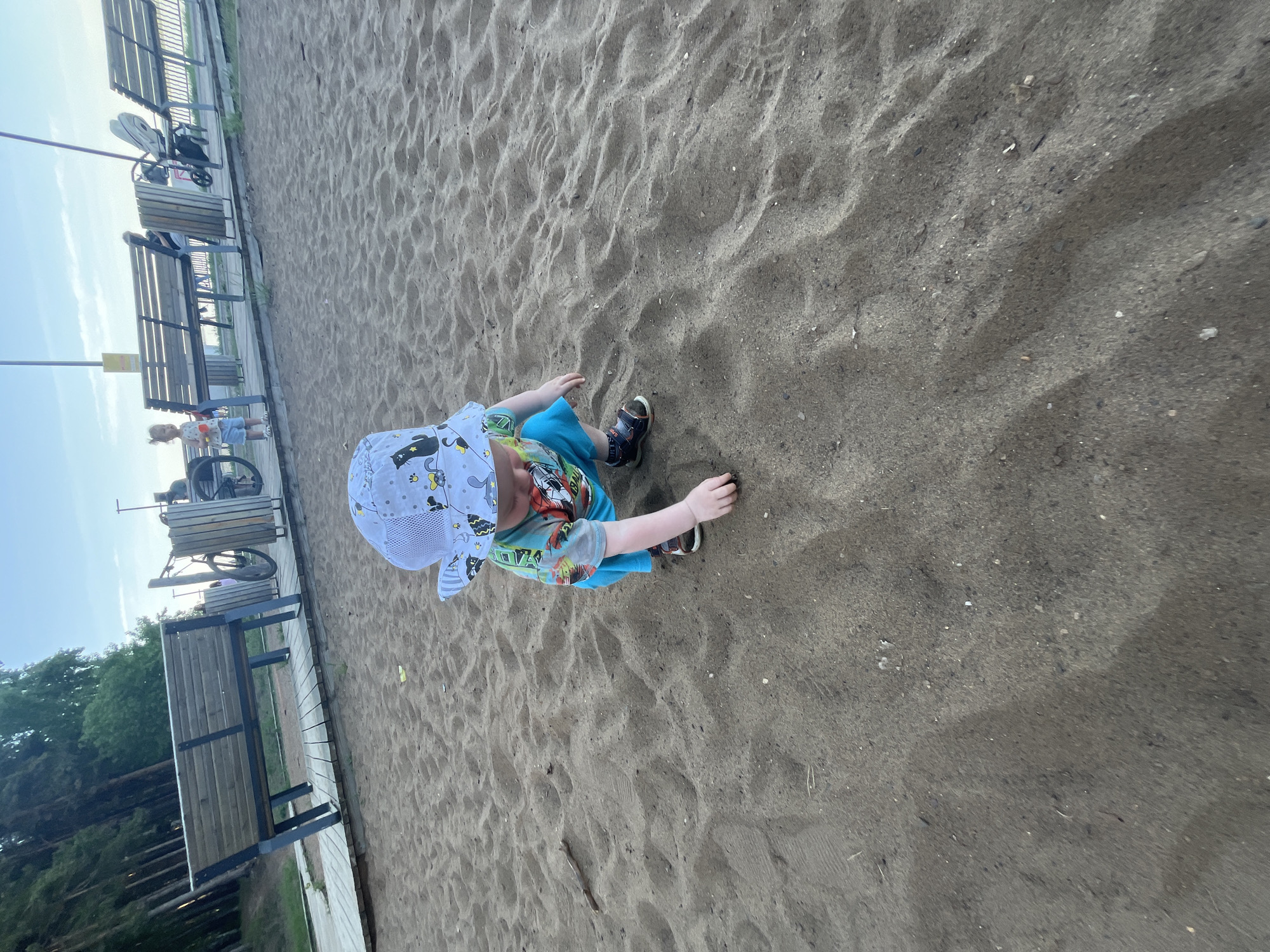 A high-angle shot captures a toddler playing in a sandy area, likely a playground, near benches. The child, wearing a patterned hat, t-shirt, and shorts, is crouched, reaching towards the sand. In the background, there are benches with metal frames and wooden slats, and a stroller is parked nearby. In the distance, another child is visible. The location is in the city of Chaikovsky, Russia. The ground is mostly sand with visible rocks.