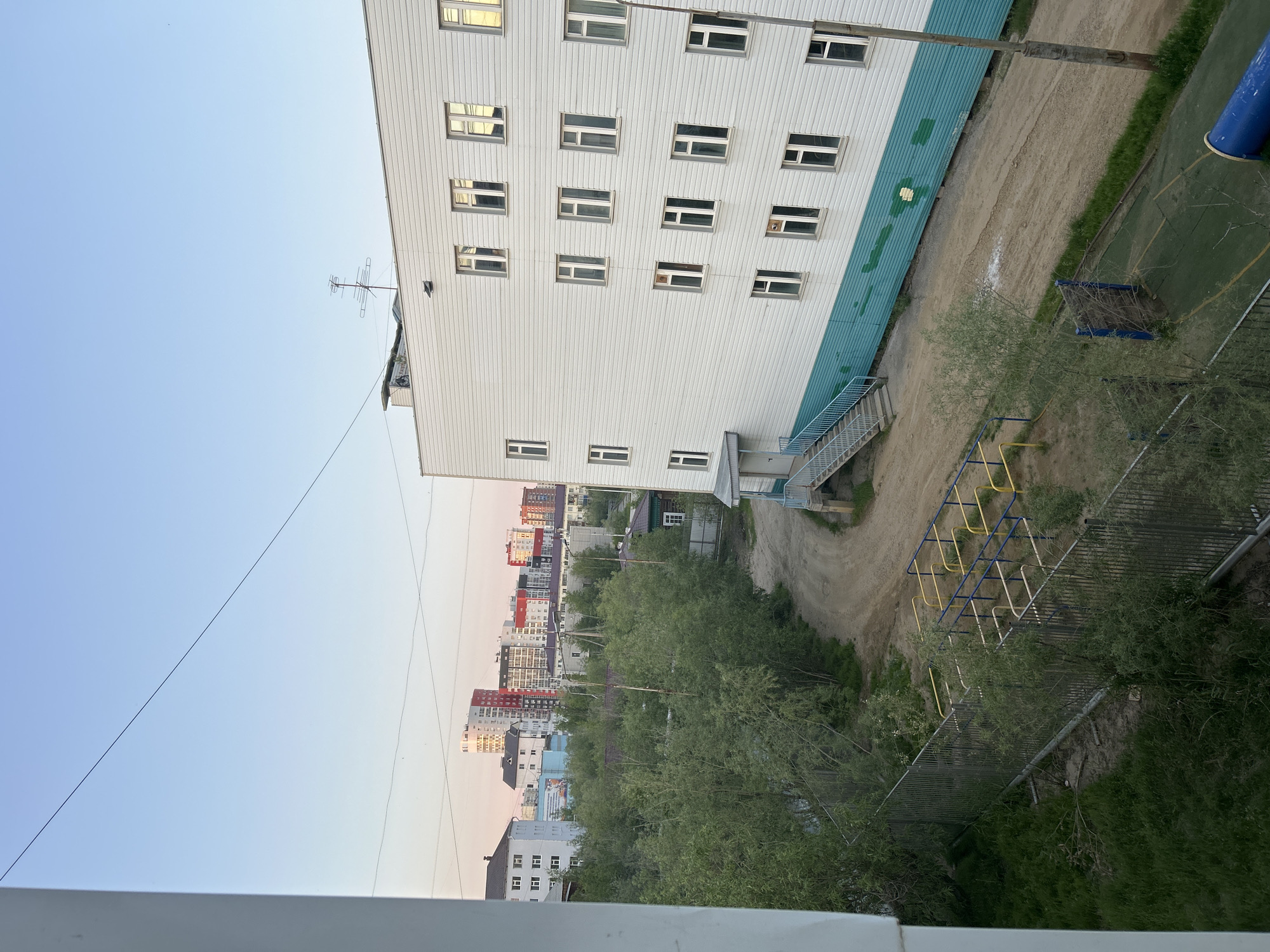 This image, taken in Yakutsk, Russia, presents an outdoor scene in what appears to be a residential area during twilight. The foreground features a section of a white building with a turquoise-green lower facade, characterized by multiple rectangular windows. To the left, a metal fence encloses an area with green grass and some vegetation. Beyond this, a dirt path winds through a sparsely treed area. Further in the background, a cluster of multi-story buildings, including some taller, modern structures with colorful facades, is visible against a pale pink and blue sky. A small playground structure with yellow and blue bars is situated near the dirt path. The overall impression is one of a suburban or outer-city environment in a northern climate, likely during the summer due to the presence of trees.