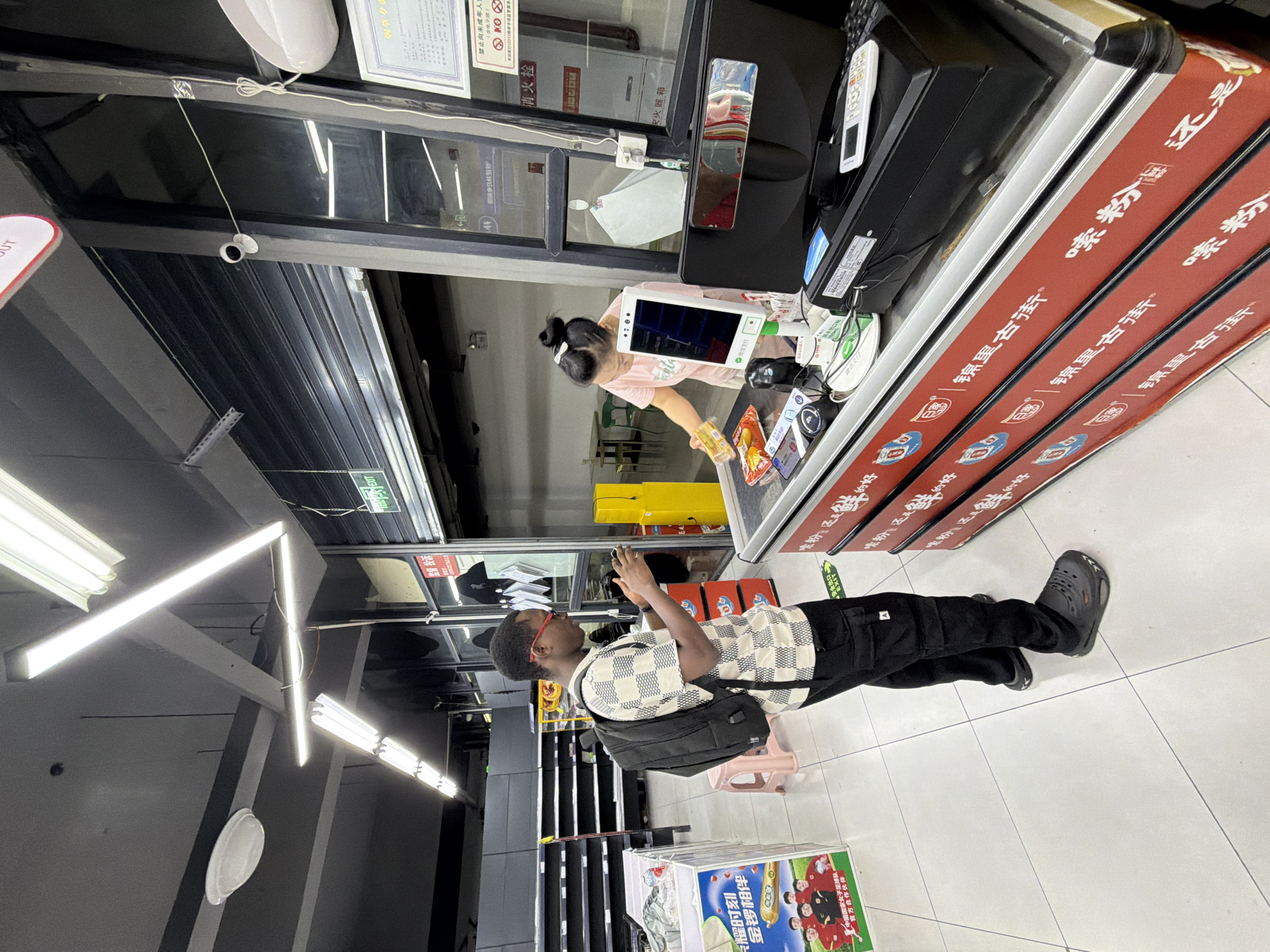 The image depicts the interior of a supermarket in Jiangjin, China, likely during daylight hours. In the foreground, a Black man wearing a checkered shirt, black cargo pants, and black crocs stands at a checkout counter, holding a mobile phone. He is wearing glasses and a backpack. Facing him is an Asian woman, presumably a cashier, wearing a pink t-shirt. She is standing behind the checkout counter, holding a yellow box and reaching for items on the conveyor belt. The checkout counter is a prominent red structure with Chinese text. Various electronic devices, including a payment terminal and a computer monitor, are visible on the counter. The background shows shelves, some of which appear empty, and fluorescent lighting fixtures on the ceiling. A security camera is also visible on the upper right side of the frame. The overall environment suggests a retail setting with customers interacting with the checkout process.