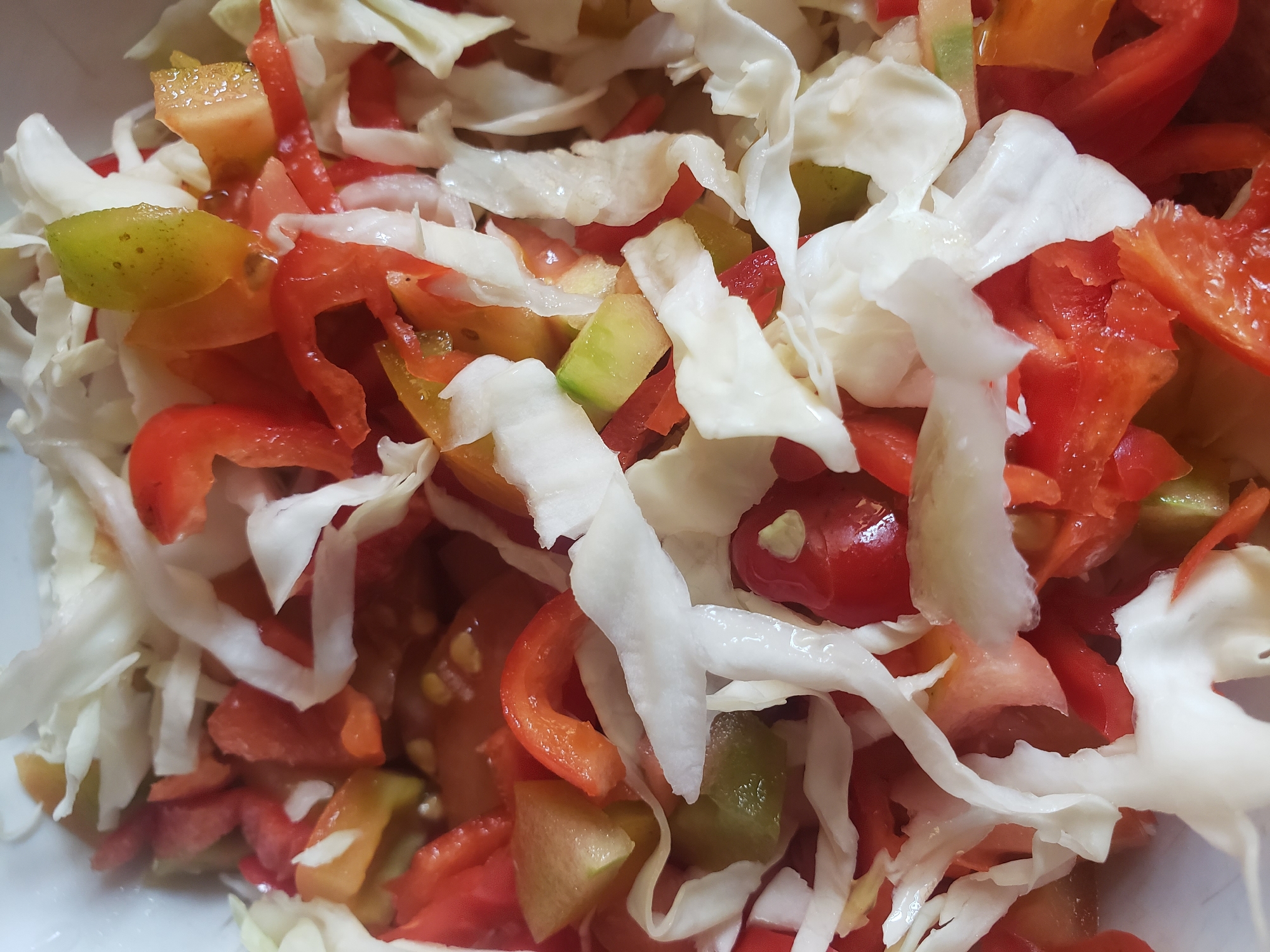 A close-up, overhead shot shows a bowl of finely chopped salad ingredients. The salad is composed primarily of shredded white cabbage, diced red tomatoes, and pieces of red and green bell peppers. The textures are varied, with the crispness of the cabbage contrasting with the softer, juicy tomatoes and peppers. The lighting is bright, highlighting the vibrant colors of the vegetables. The image does not provide any information about the city of Narayi, Nigeria, or any specific environmental context.