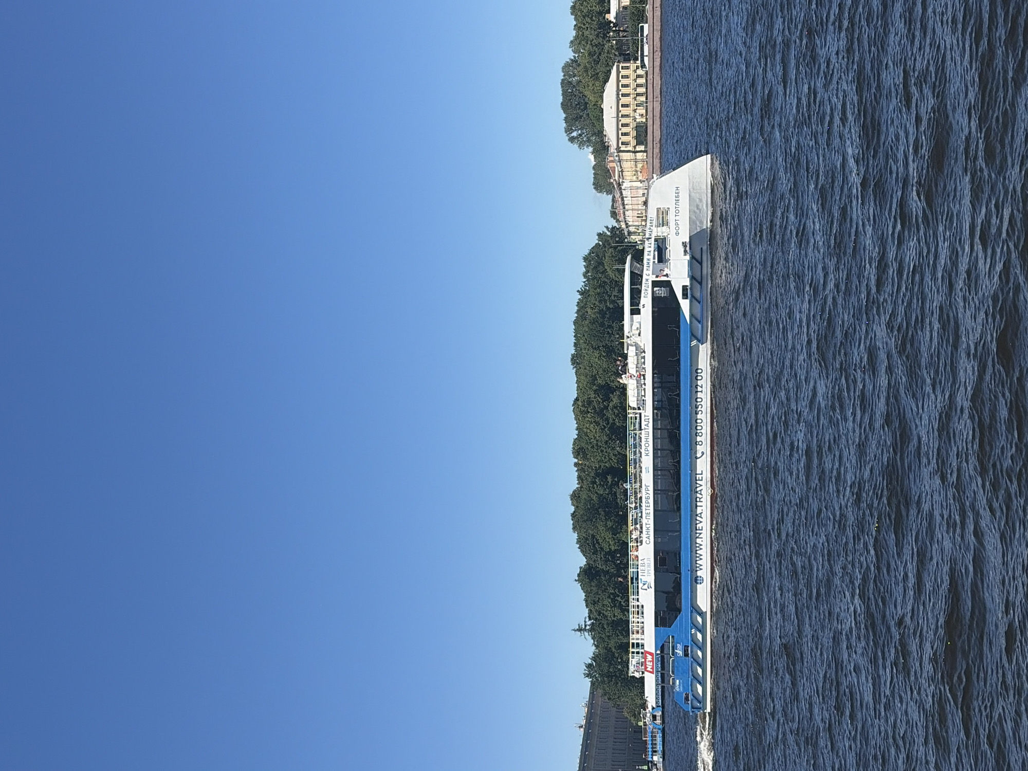 A white and blue passenger boat is cruising on a dark blue body of water under a clear, bright blue sky. The boat has "Neva Travel" written on its side in Russian and English, along with a phone number and website. There are passengers visible on the upper deck. In the background, a lush green tree line runs along the shore, with several buildings visible behind the trees, including a light yellow building with arched windows. The scene appears to be in Saint Petersburg, Russia, likely along the Neva River or a canal, given the presence of the "Neva Travel" boat and the architecture.