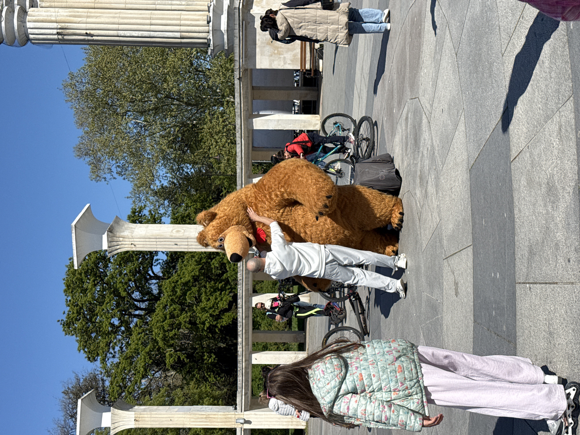 The image shows a sunny outdoor scene in Varna, Bulgaria, specifically at the Pantheon. A large brown bear mascot is positioned near the edge of the raised area, with a person in a white tracksuit interacting with it. In the background, several people are walking or riding bicycles along the pavement, and others are seated on benches. Classical columns and trees are visible, defining the Pantheon's architectural style. The ground is paved with large gray stone slabs.