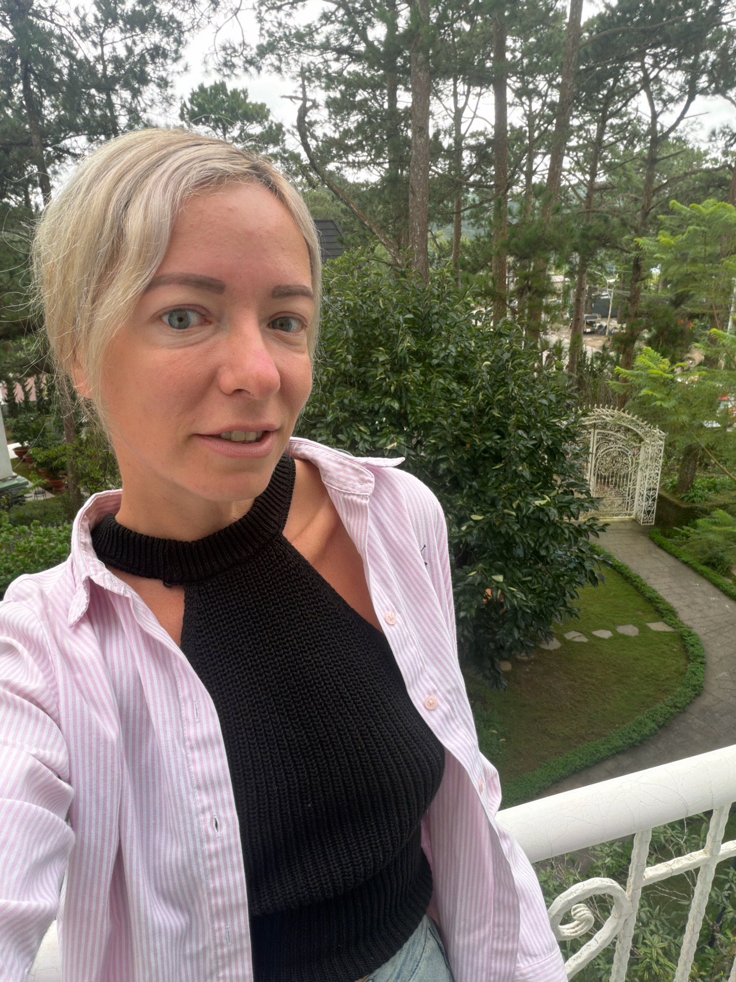 In the city of Xuân Trường, Đà Lạt, Vietnam, a woman with short blonde hair stands on a balcony, taking a selfie. She is wearing a pink and white striped button-down shirt, open to reveal a black knitted top underneath. Her gaze is directed at the camera, and her mouth is slightly open. Behind her, a lush green landscape unfolds, featuring tall pine trees, manicured gardens with a winding path, and an ornate white metal gate. The sky is overcast.