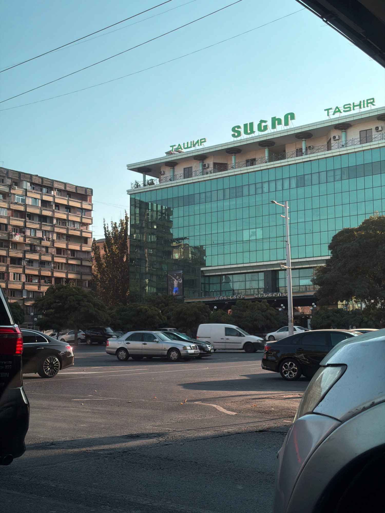 This is a medium-shot, eye-level outdoor scene in Yerevan, Armenia, focusing on a street with several cars and a modern glass-fronted building. The sky is clear and blue, with overhead power lines visible.
In the foreground, the rear of a silver car is partially visible, obscuring the left side of the frame. To the left, the rear of a black car is also visible. Beyond these, a silver Mercedes-Benz C-Class is parked on the right side of the street, and further back, a white van and a dark-colored sedan are parked. A multi-story apartment building, typical of Soviet-era architecture, with numerous balconies, stands to the left of the main building.
The central focus is a multi-story building with a facade made of reflective, greenish-blue glass. The building has a distinct upper level with a white facade and decorative columns, topped with green signage. The signage includes the word "TASHIR" in English and what appears to be Armenian script. The building also features air conditioning units visible on the upper levels. A large advertisement featuring a man in athletic wear is displayed on the lower part of the glass facade. Trees and dense shrubbery are situated in front of the building, and a lamppost stands in front of the glass facade, reflecting in the glass. The road surface is asphalt with visible painted lines. The time of day appears to be late afternoon, with long shadows cast across the road.