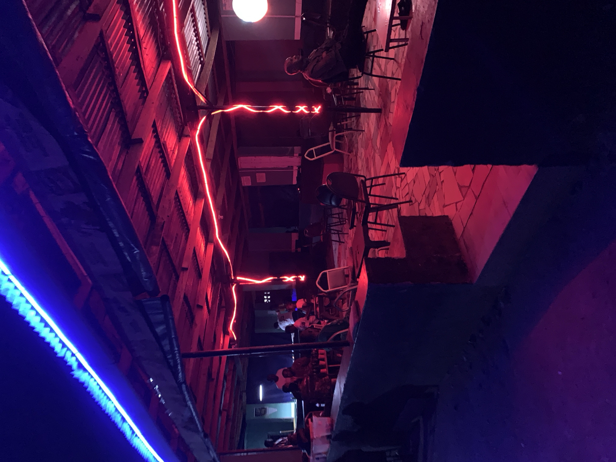 Captured at night near New Stadium in Nigeria, the scene reveals an outdoor space with corrugated metal walls illuminated by red and blue neon lights. The floor is paved with tiles, and several tables and chairs are arranged, occupied by individuals, suggesting a social gathering. Overhead, a single white light bulb provides additional illumination. The overall ambiance is dimly lit and visually striking due to the contrasting neon colors.