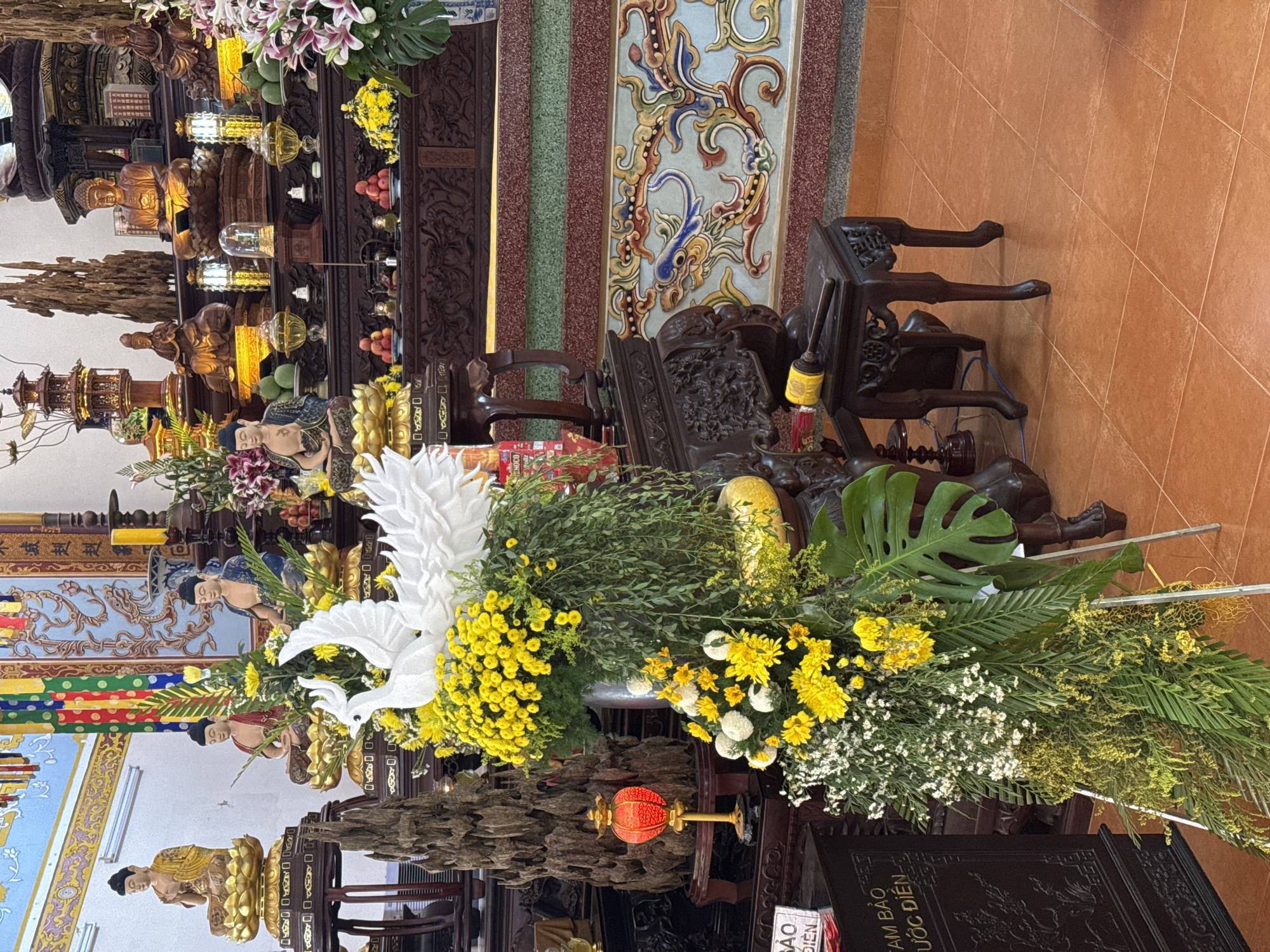 The image shows the interior of what appears to be a Buddhist temple or shrine in Nha Trang Ward, Vietnam. The foreground features a floral arrangement consisting of white doves, yellow flowers, greenery, and a red lantern. The arrangement sits on a carved wooden stand. Behind it, there is a series of tiered wooden structures holding statues, offerings, and religious objects. The statues appear to be of Buddha and other figures associated with Buddhism. The background includes decorative wall panels with ornate patterns and colors. The floor is covered in terracotta tiles. A dark wood chair is visible to the right.