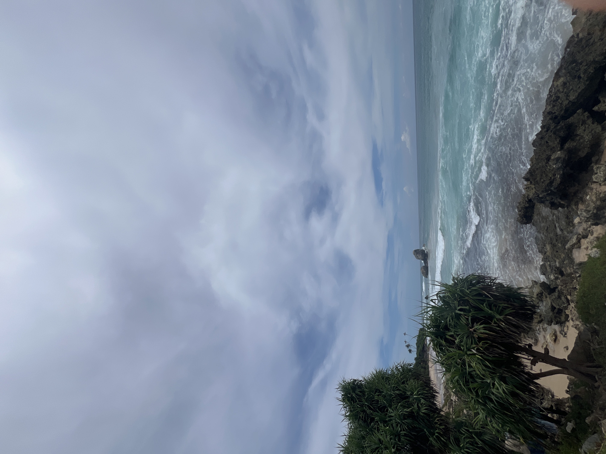 The image shows a view of the beach at Kathaluwa, Sri Lanka. The ocean waves are crashing on a rocky shoreline. The sky is cloudy. There are trees in the foreground.
