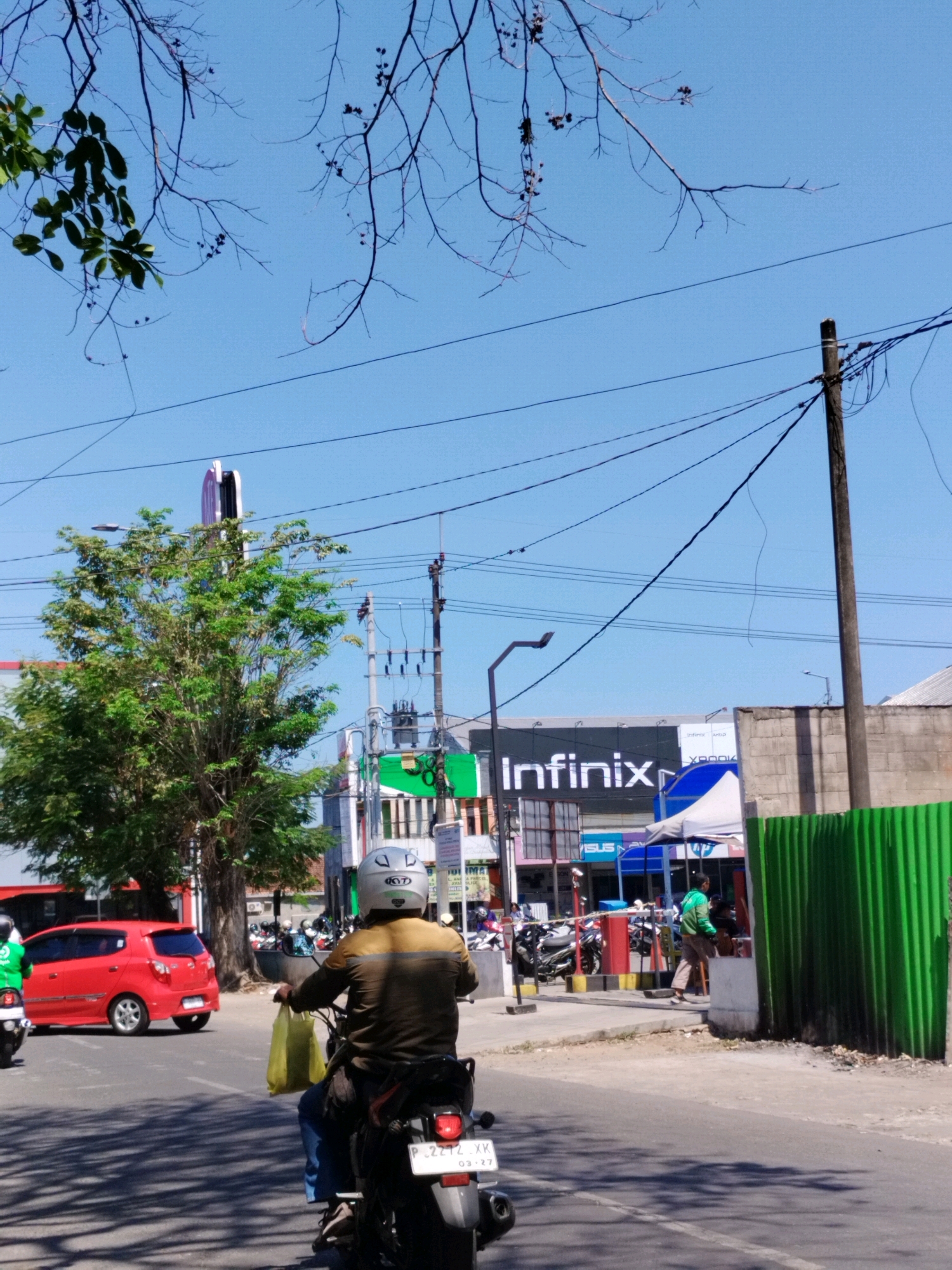In Jember, Indonesia, a street scene unfolds under a clear blue sky. In the foreground, a person on a motorcycle, wearing a light brown jacket and a white helmet, rides away from the camera. The motorcycle has a license plate that reads "P 2272 XKK". The rider is carrying a yellow bag on their left side. To the left of the motorcycle, a red car is parked or moving slowly. A large tree with green leaves stands on the left side of the road, partially obscuring a tall, illuminated sign with a curved top. Further down the road, a building displays a large "Infinix" advertisement. Power lines crisscross the sky, supported by wooden and metal poles. A green corrugated metal fence runs along the right side of the road. Several other motorcycles are visible parked or moving in the background. The overall impression is a typical day in a bustling Indonesian city.
