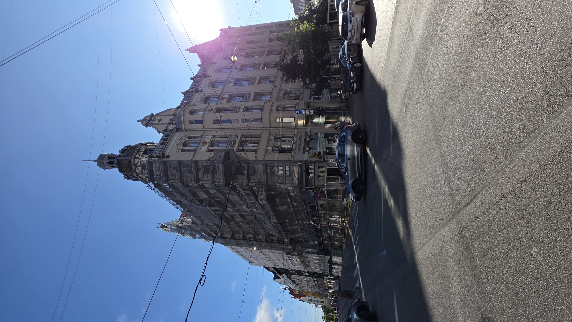 The image depicts a street scene in Riga, Latvia, focusing on a large, ornate building undergoing renovation, indicated by extensive scaffolding and netting covering its facade. The building is situated at a street corner with a distinctive tower. Several cars are visible on the paved road, including a dark grey SUV in the foreground and other vehicles further down the street. A pedestrian can be seen crossing the street at a marked crosswalk. The sky is clear blue with a bright sun casting strong shadows across the road and the building. The architecture suggests a European city. The presence of a "P" sign indicates parking is available nearby.
