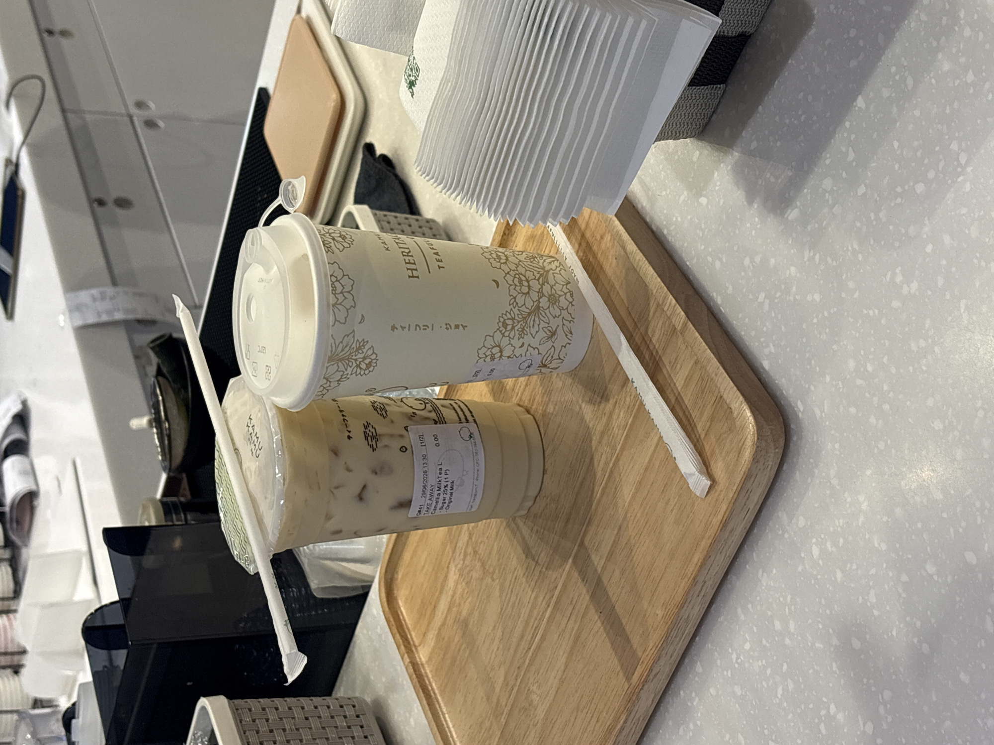 The image captures a close-up view of a counter in a beverage establishment, likely in Nong Pa Khrang, Thailand. Two takeaway cups are prominently displayed on a wooden tray. The cup on the left, a clear plastic one, contains a light-colored milky beverage with ice, and its label indicates "Camellia Milk Tea L, Sugar 25% (1 P), Original Milk." A white paper straw is resting on its lid. The cup on the right is made of cream-colored paper, adorned with floral patterns and text that appears to be in Japanese and English, reading "KAHO HERITAGE TEAFLOWER." Another white paper straw is positioned on its lid. To the right of the cups, a stack of folded white paper napkins is visible in a grey woven basket. The counter surface is light grey with a speckled texture. In the background, a black dispenser unit and other indistinct objects are partially visible. The overall lighting suggests an indoor environment.