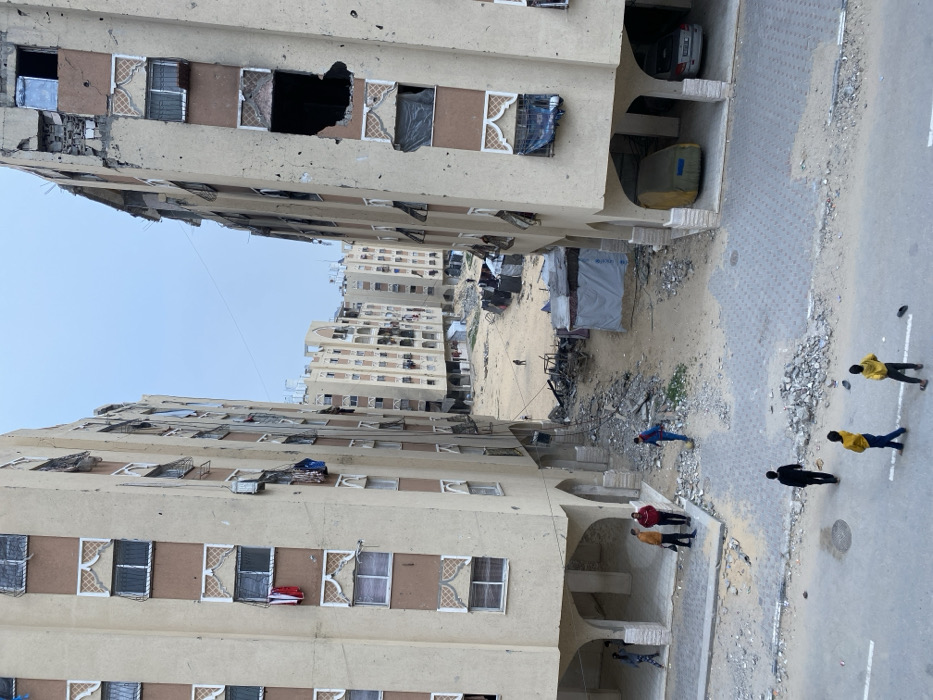 This image depicts a damaged residential area in Khan Yunis, Palestinian Territory, characterized by extensive structural damage to multi-story apartment buildings.
In the foreground, a paved road with scattered rubble transitions into a sandy, debris-strewn open area flanked by two tall, beige-colored apartment blocks. These buildings feature multiple floors with decorative window frames, many windows shattered, missing, or boarded up. The building on the right exhibits particularly severe damage, including large, irregularly shaped holes in its upper-story facade. Laundry hangs from several windows on both buildings.
Under the ground floors of these buildings, which are elevated on pillars, are covered spaces. On the left, several males stand and converse, while another individual walks past. On the right, two vehicles are parked: one covered in a yellow tarp, and another silver sedan. A male figure in a blue shirt walks across the rubble near the right building's base.
The central open space between the buildings is primarily sand and dirt, littered with large piles of rubble, twisted metal, and what appears to be burnt wreckage. Several makeshift shelters, constructed from tarps and corrugated metal, are visible in this area, including one structure covered by a blue tarp. In the distance, numerous other multi-story residential buildings, also appearing damaged, line the horizon. A lone figure walks across the central sandy expanse further into the scene.
In the immediate foreground, on the paved road, three young individuals are walking away from the viewer; two are wearing yellow shirts, and one a dark shirt. The sky is overcast and light gray.