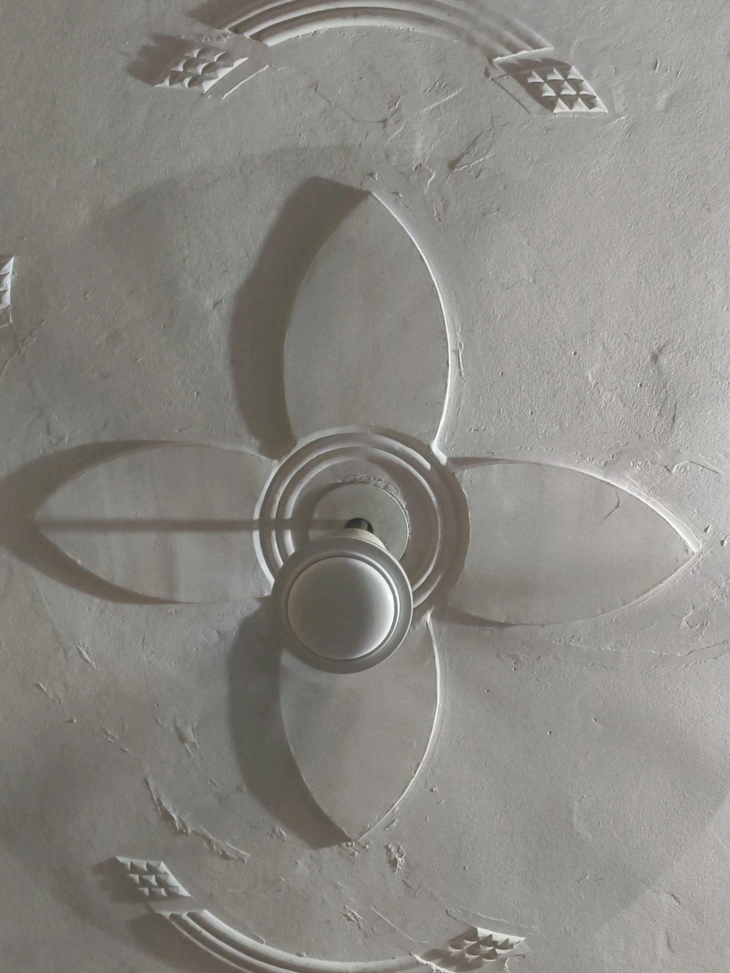 The image shows a ceiling fan with a decorative plaster design. The fan is centrally located, with its blades casting shadows on the ceiling. The plasterwork features a floral motif with four petal-like shapes radiating from the center. There are also geometric patterns on the upper and lower portions of the ceiling. The ceiling appears to be made of concrete and is painted white. The lighting suggests an indoor environment. The image does not provide any information about its location in Pura, India, or any details about people, actions, or interactions.