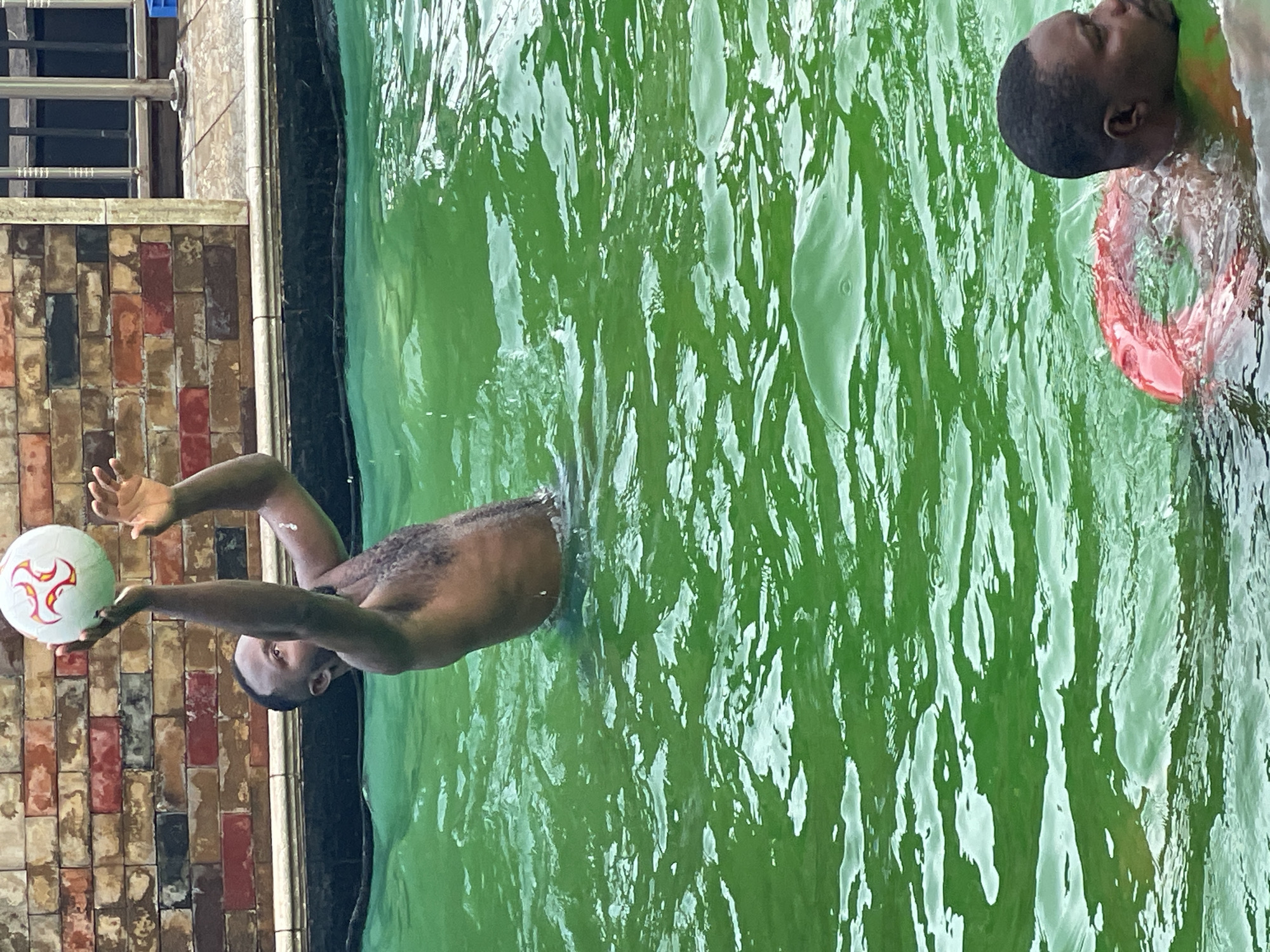 In an outdoor swimming pool with green water, two Black men are visible. The man in the foreground is partially submerged and looking to his right. He is wearing a red flotation device around his torso. The second man, further in the background, is also partially submerged and is in the process of tossing a white and red soccer ball upwards with his hands raised. Behind the pool, a brick wall with multi-colored bricks forms the background, with a metal fence and tiled area visible to the right.
