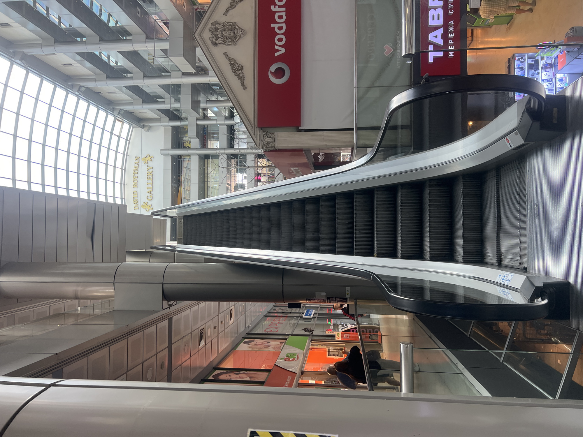 The image depicts the interior of a multi-story shopping mall, likely in Odesa, Ukraine, given the context provided. A prominent escalator, with its characteristic ribbed steps and black handrail, occupies the foreground, ascending towards the upper levels of the building. The mall's architecture features a large, arched glass ceiling that allows natural light to filter in, illuminating the various levels and shops.
Several shops are visible on different floors. On a lower level, to the left of the escalator, there are storefronts with large displays. One prominent display features images of women, with signage that appears to be in Cyrillic. Another shop has a green and orange logo that reads "eva". Further up, a large red sign with a white circular logo and the text "vodafone" is visible, indicating a telecommunications store. Above this, a sign reads "DAVID ROYTMAN GALLERY" in yellow lettering.
There are a few people visible in the scene. In the lower left, a person wearing a dark top and light-colored pants is walking away from the camera, and another person is partially visible behind them. On the same level as the "eva" store, a man in a dark jacket with red accents and a face mask is standing, seemingly interacting with a display. Further in the background, on an upper level, other individuals can be observed moving within the mall.
The environment appears clean and modern, with polished floors and glass railings separating the different walkways and balconies. The overall impression is of a busy commercial space with multiple retail outlets and a constant flow of shoppers.