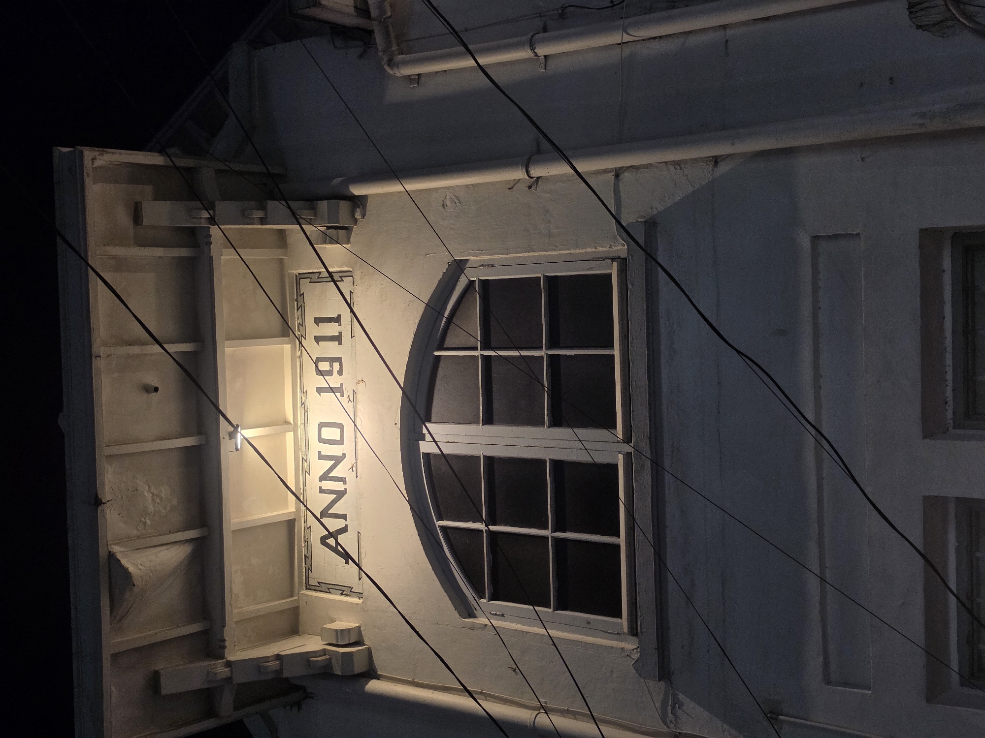 The image shows the facade of an old building at night, illuminated by a spotlight. The building has a sign that reads "ANNO 1911" above a large arched window with multiple panes. Several dark power lines are strung across the facade. The building appears to be made of light-colored brick or stucco, with some decorative architectural elements. The overall impression is one of age and historical significance.