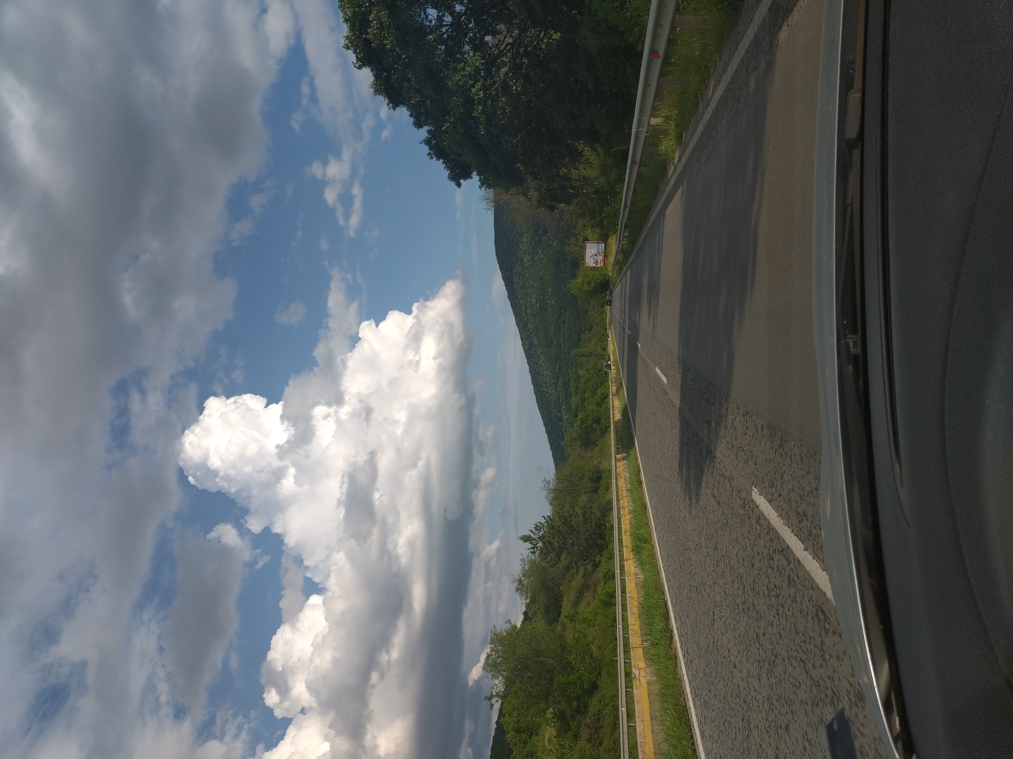 The image depicts a straight asphalt road cutting through a verdant, hilly landscape, viewed from the interior of a vehicle near Zmeyovo, Bulgaria. The sky above is partially covered by a mix of bright white cumulus clouds and darker grey stratocumulus clouds, with patches of clear blue sky visible. The two-lane road features a white solid line marking the left edge and a dashed white line for lane separation further ahead.
On the left side of the road, a yellow and white concrete barrier topped with a metal guardrail runs parallel to the pavement. Beyond this barrier, a steep, densely forested hillside rises, covered with various shades of green deciduous trees. On the right side, a metal guardrail separates the road from a grassy verge and a large, mature tree with dark green foliage. Further down the road on the right, another tree-covered hill is visible, and a billboard displaying an advertisement stands beside the road.
Several vehicles are observable in the distance on the road, traveling in both directions. The scene is illuminated by natural daylight, casting shadows on the road surface, suggesting a bright, partly sunny condition. The dashboard and a portion of the windshield are visible in the foreground, indicating the perspective from inside a car. No individuals are explicitly identifiable.