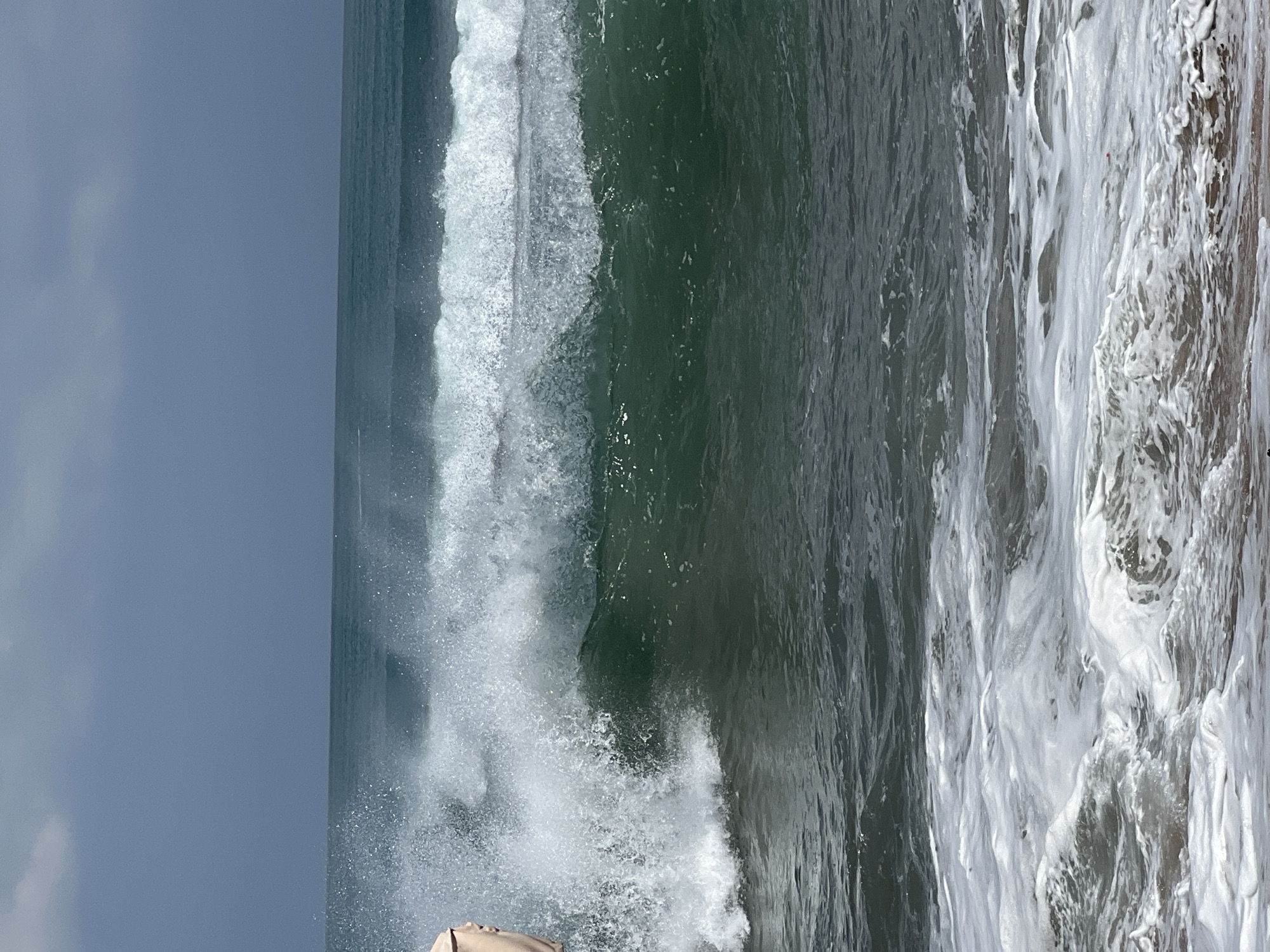 A vertical, medium shot captures a breaking wave at Kuta Beach, Indonesia. The ocean is dark green and turbulent, with white foam churning around the wave's crest. The sky above is a muted grey, with hints of clouds. On the far left of the frame, a person's arm and shoulder are visible, possibly indicating they are standing near the water's edge or have just been splashed. The foreground shows the sandy shore with incoming foamy water.