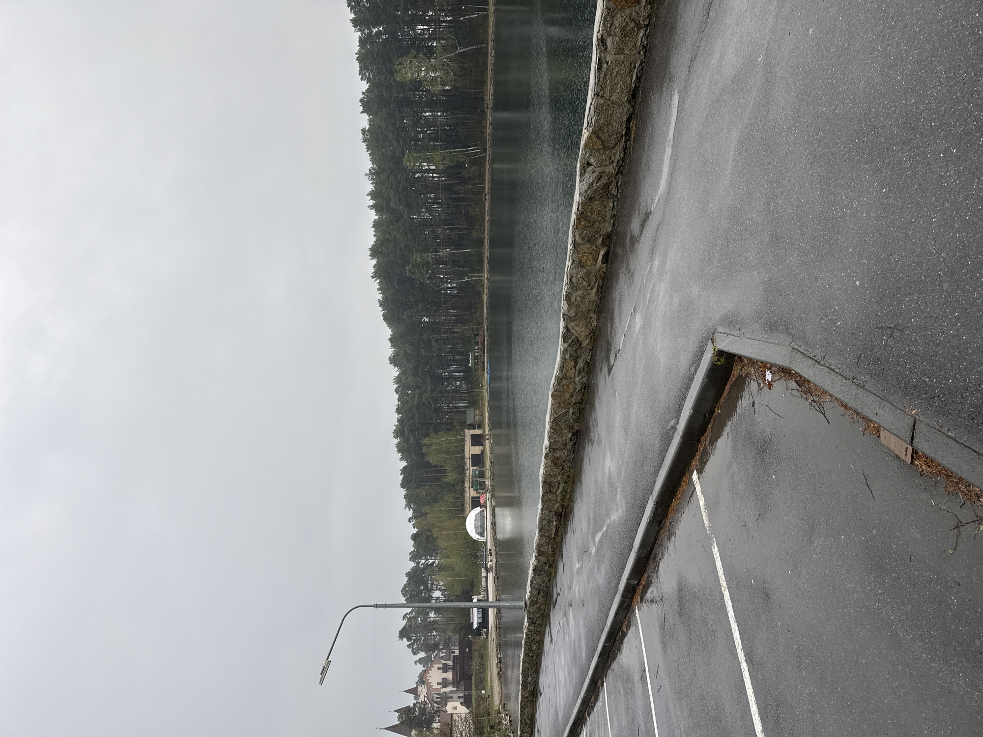 The image depicts a lakeside scene in Turgoyak, Russia on a rainy day. A wet parking lot with white painted lines sits in the foreground. A stone retaining wall separates the parking lot from the lake. Across the lake, a dense forest line is visible. Buildings and a white domed structure are situated along the opposite shore. A street lamp is also visible on the left side of the frame.