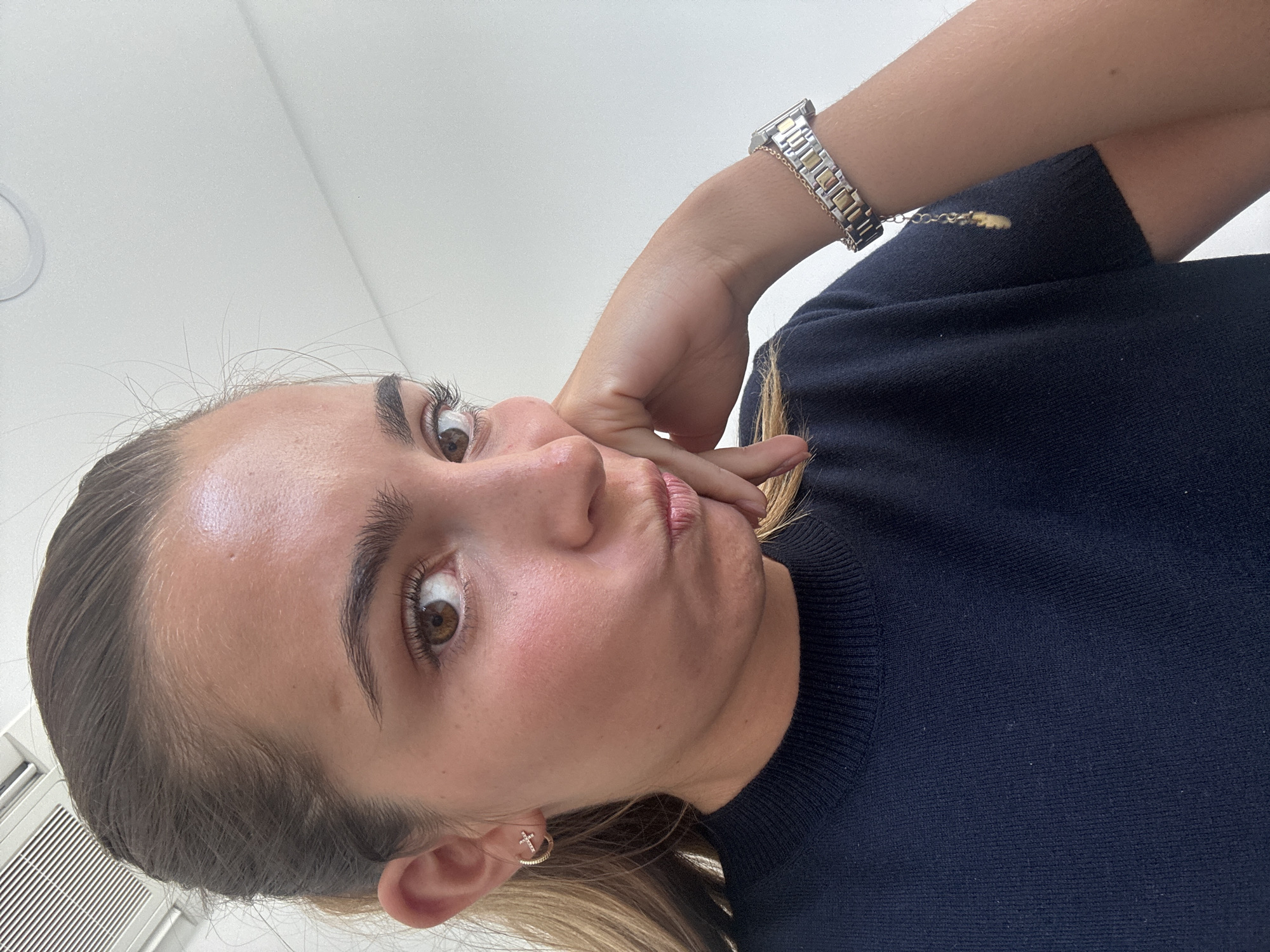 A light-skinned woman with brown hair and eyes is visible in a close-up, slightly distorted photograph, taken from a low angle. She is wearing a dark blue sweater and a silver and gold wristwatch on her left wrist. She is holding her hand to her face, resting her fingers against her cheek and lips. The background is a plain white wall. The woman has a small gold cross earring in her left ear. The photograph does not contain any location information about Torres Vedras, Portugal.