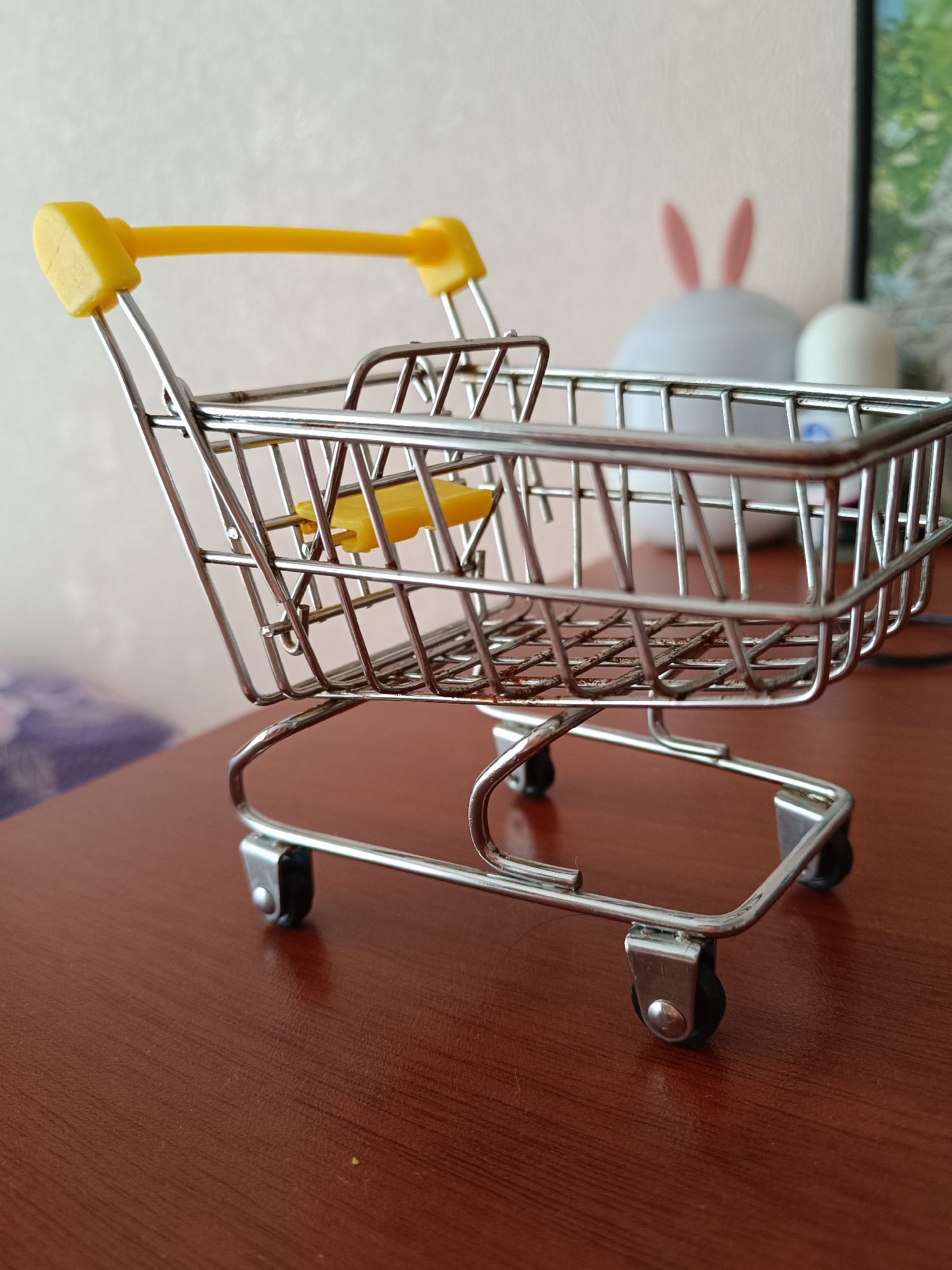A miniature metal shopping cart with yellow plastic handles rests on a brown, wood-grain surface. In the blurred background, a white lamp with pink bunny ears is visible, along with part of a framed picture. The cart's wireframe construction and small black wheels are clearly detailed. The image appears to be taken indoors. The location is not discernible from the image.