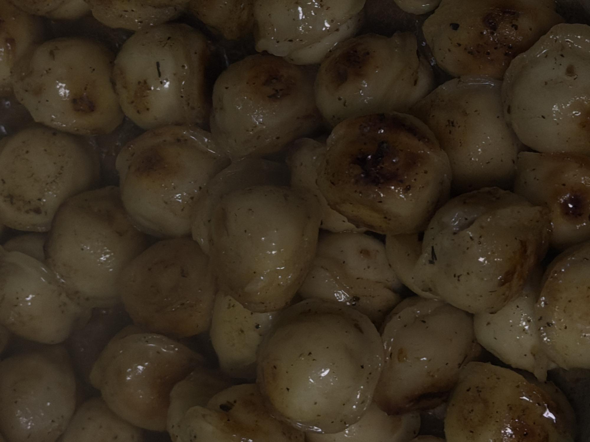 The image is a close-up, overhead shot of a pile of gnocchi or dumplings. The food is golden brown with some darker caramelized spots, indicating it has been cooked, likely pan-fried or roasted. Each piece is round and plump, coated in a glossy sauce or oil, and speckled with small dark flecks, possibly herbs or spices. The lighting is dim, creating a warm and slightly moody atmosphere. The image does not contain any discernible details related to the city of Bobrovo, Russia, or any environmental or human elements.