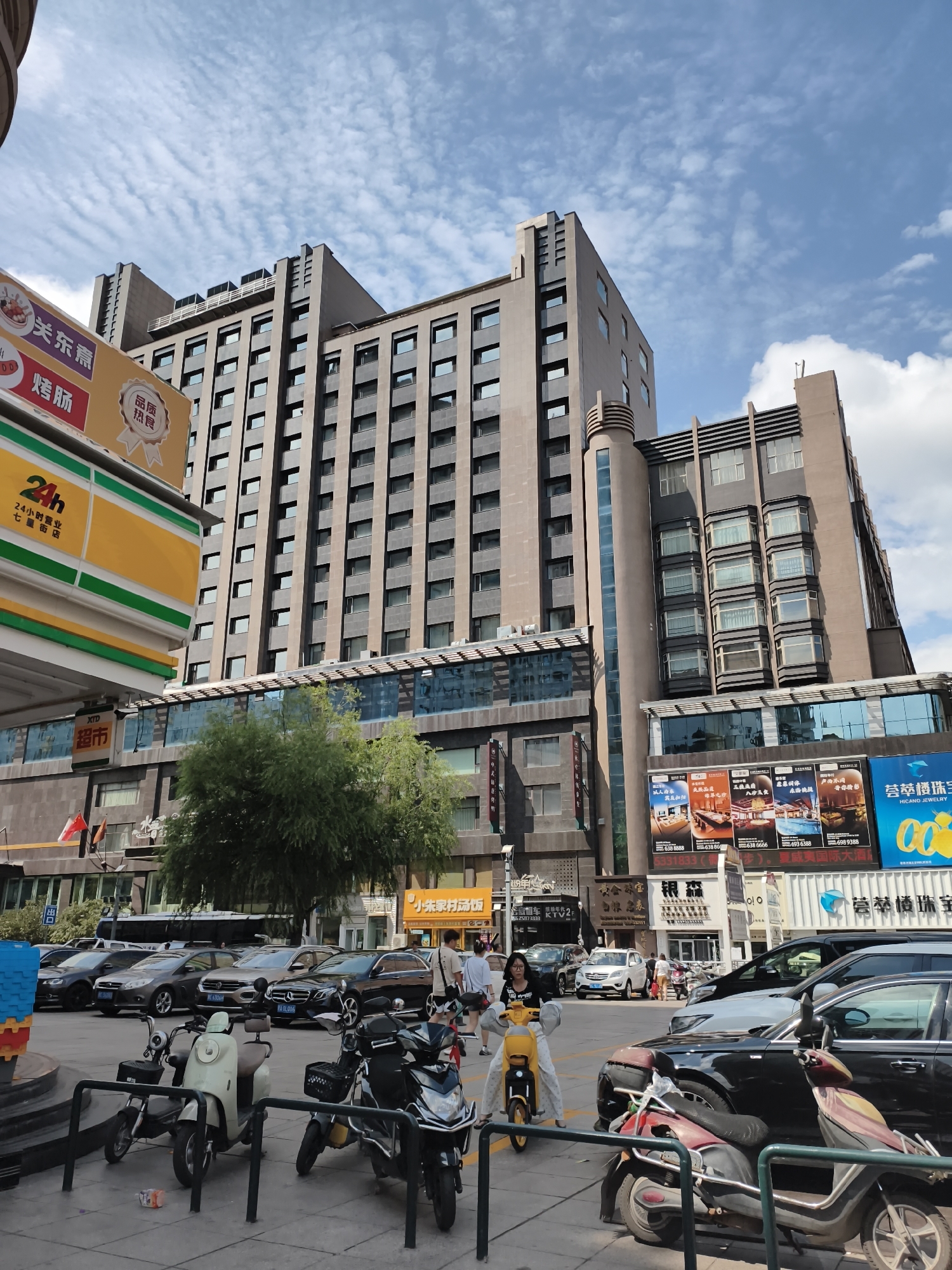 A wide shot captures a street scene in Mudanjiang, China, dominated by a modern, multi-story building with a dark brown facade. The building features numerous rectangular windows and decorative architectural elements, including vertical lines and stepped sections. In front of the building, a row of cars is parked on asphalt. Several motorcycles and scooters are also parked, with three prominent in the foreground. A woman is seated on a yellow electric scooter in the mid-ground. Other pedestrians are visible walking in the background. A large sign with Chinese characters and images of food is on the left side of the frame, part of what appears to be a convenience store with a "24h" logo. Several other advertisements with images and Chinese text are displayed on the building's lower levels. The sky above is blue with scattered white clouds. A large tree with green foliage is situated in front of the building, partially obscuring the lower floors.