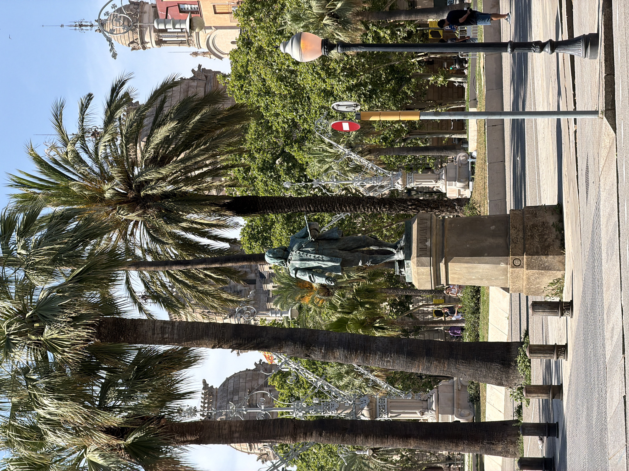 The image depicts a public square in Barcelona, Spain, likely Passeig de Gràcia due to the architectural style of the buildings in the background and the presence of palm trees and decorative lampposts. A bronze statue of a man, identified by the inscription "VILADOMAT" on its pedestal, stands prominently in the center of the frame. The statue depicts a man in historical attire, holding a tool, possibly a brush or a chisel, suggesting he is an artist or craftsman.
In the foreground, several dark, cylindrical bollards are arranged in a line on a paved walkway. Behind the statue, a lush green area with more palm trees and dense foliage is visible, with ornate, wrought-iron street lamps adding to the aesthetic. In the background, the distinctive architecture of Barcelona is evident, with ornate building facades and towers suggesting a historic urban setting.
Several people are present in the scene, though they are small and somewhat indistinct. Two individuals are seen walking in the background, near the green area, and another man in a dark t-shirt and blue shorts stands near a street lamp on the right side of the image. The sky is clear and blue, indicating a sunny day. The overall scene conveys a sense of urban tranquility and cultural heritage.
