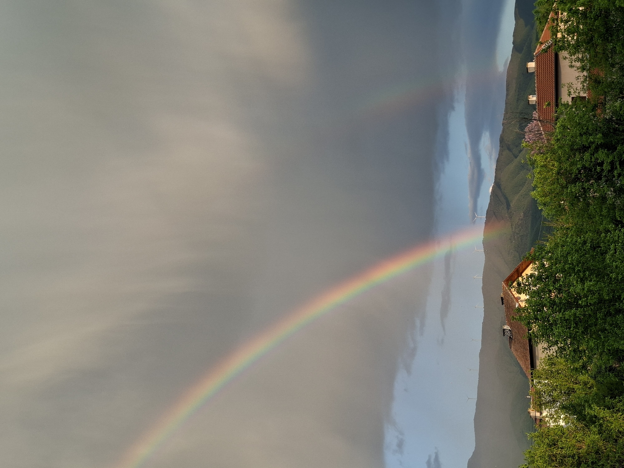The image shows a landscape featuring a rainbow. The rainbow's arc spans from the left side of the image towards the center, appearing against a backdrop of cloudy skies. On the right side, the landscape transitions to reveal mountainous terrain, partially obscured by trees and shrubbery in the foreground. A building with a red-tiled roof is visible amidst the greenery. Further in the background, wind turbines stand on the mountain ridge. The city is Unknown, Unknown.