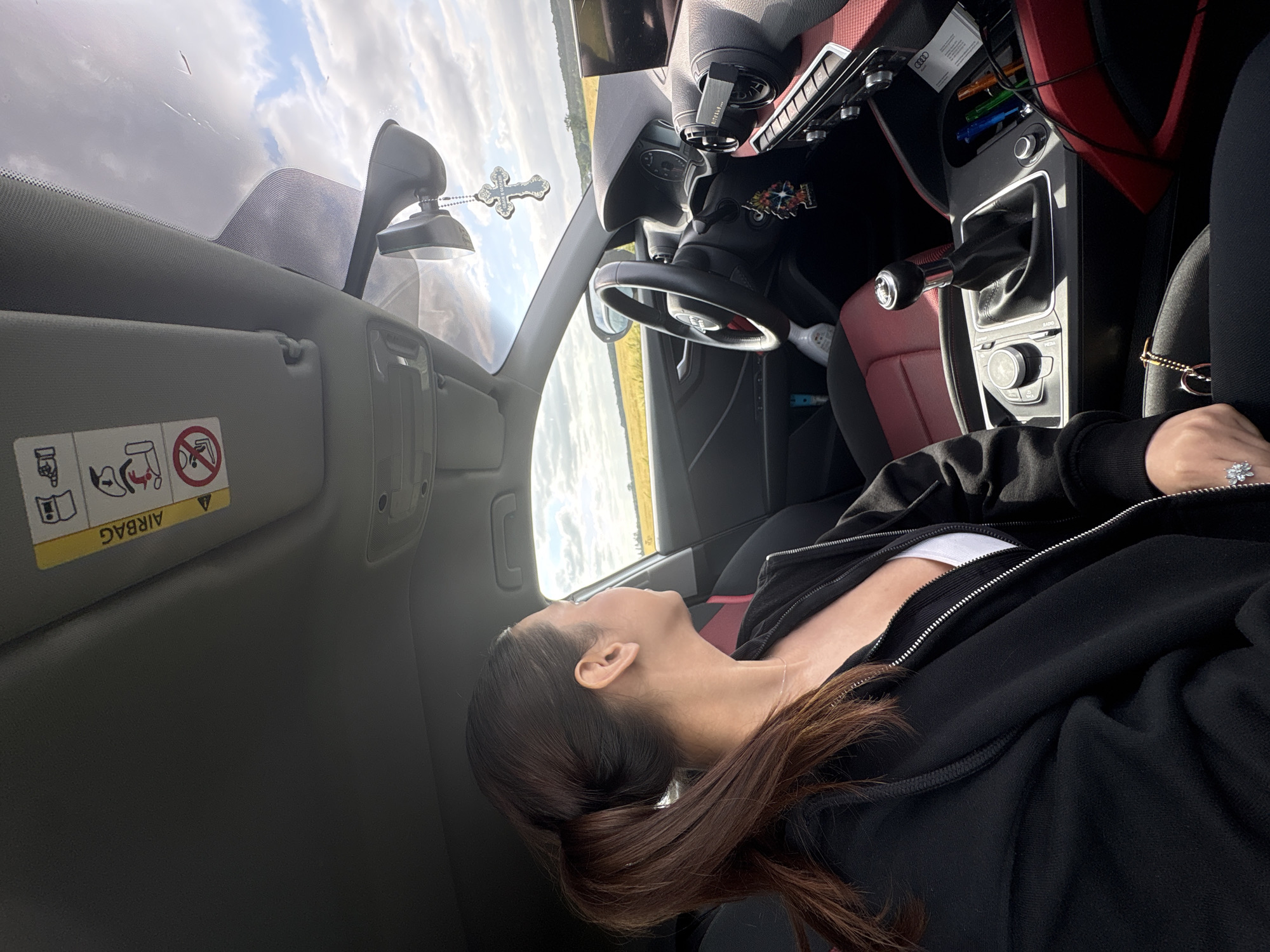 An interior view of an Audi vehicle, captured from the perspective of the front passenger seat, shows a single adult female in the foreground, seated in the passenger seat and looking out the window towards the driver's side. The scene is located in Wegberg, Germany.
The female has dark brown hair styled in a ponytail and is wearing a black zippered hoodie over a white top, along with a delicate necklace. A prominent ring with a clear stone is visible on her right ring finger. Her left arm and hand rest on her lap.
The car's interior features a black dashboard with red accents on the door panels and center console. The steering wheel, partially visible on the left, bears the Audi logo. A manual gear stick is prominently located in the center console, alongside the Audi MMI control knob and various buttons. A small white bottle, possibly hand sanitizer, is positioned near the driver's seat. A compartment in the center console holds multiple colored pens.
Hanging from the rearview mirror is a dark, intricately designed cross ornament, which appears to have text or symbols inscribed, possibly "SLAVA UKRAINI". The passenger-side sun visor is raised, displaying a yellow "AIRBAG" warning label with multiple illustrative symbols.
Outside the vehicle, a partially cloudy sky with distinct white cumulus clouds is visible. The landscape consists of a wide, open field with yellowish-brown vegetation, extending towards the horizon. The lighting suggests a bright, daytime setting.