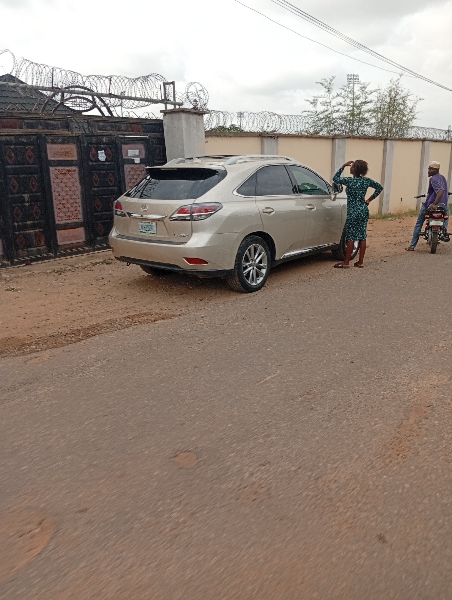 The image shows a residential street scene in Lokoja, Nigeria. A beige Lexus RX SUV is parked on the side of the road, partially blocking the pavement. A woman in a green patterned dress stands next to the driver's side of the car, her right hand touching her head, as if in contemplation or conversation. Behind her, a man wearing a purple shirt and a white cap sits on a motorcycle. In the background, there's a high wall topped with barbed wire, securing a property. Behind the car, a gate and another segment of the wall feature similar security measures. The street is asphalt, showing signs of wear and tear. The sky is overcast.