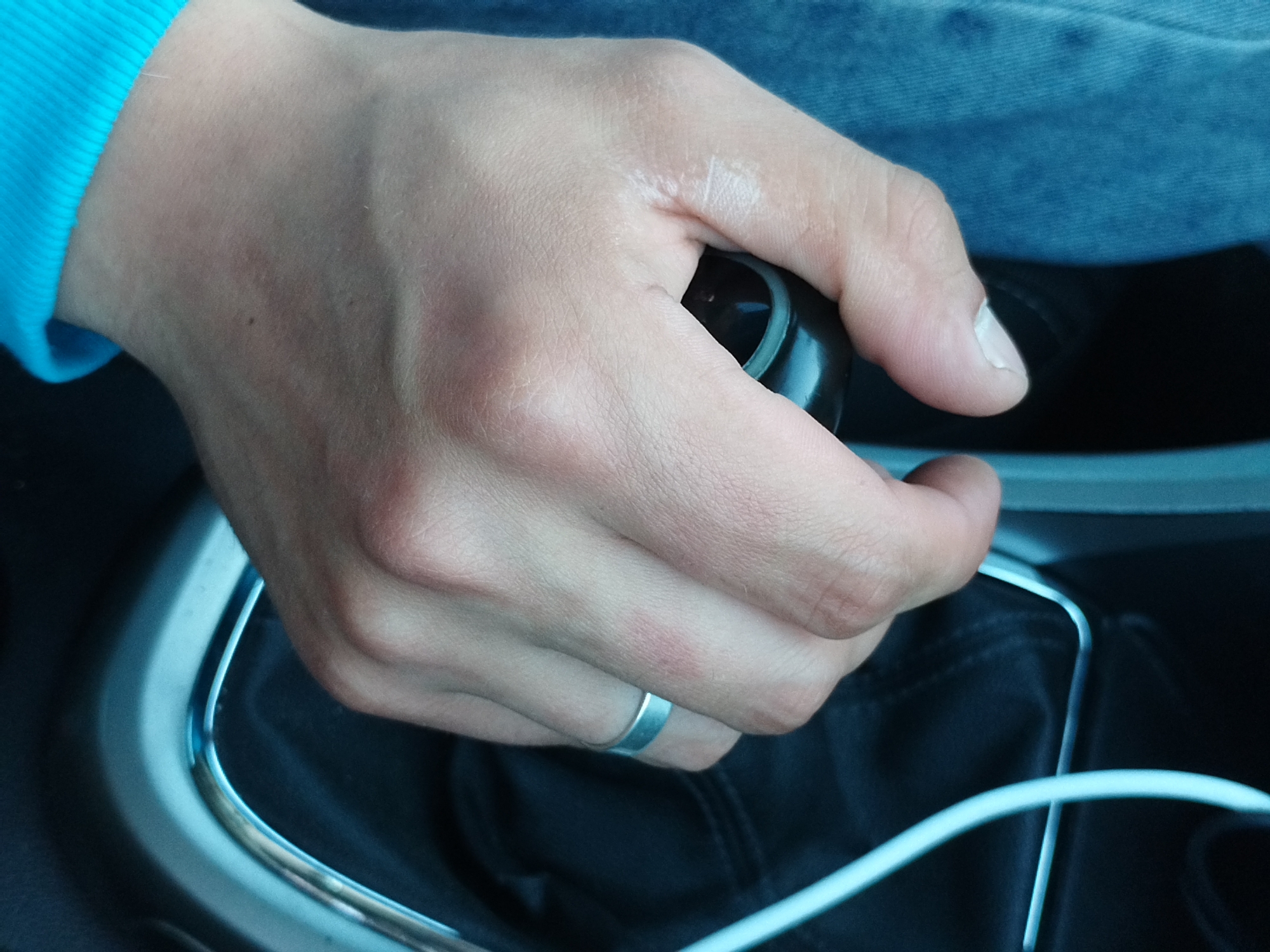 A close-up shot shows a person's hand gripping a black gear shift knob in a car. The hand is wearing a silver ring on the ring finger. The person is wearing a turquoise long-sleeved shirt. The background shows the interior of a car, including black upholstery and a blue fabric.
