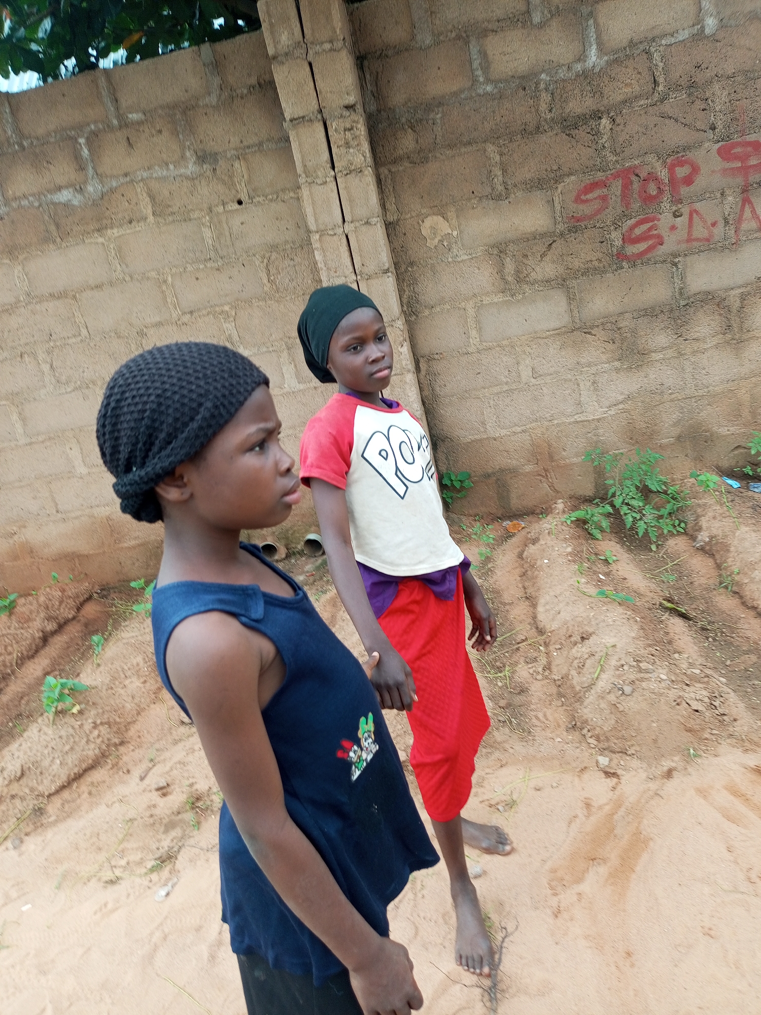 Two young girls stand on a sandy surface in front of a brick wall. The girl on the left, wearing a dark blue sleeveless dress over black pants and a black knitted cap, is looking to her left. The girl on the right, wearing a white t-shirt with red sleeves and a purple hem, a purple skirt, and a black headscarf, is looking to her right. The brick wall behind them is covered in red spray paint graffiti. The ground is composed of sand, with some sparse green vegetation growing in patches. The overall impression is of an outdoor, possibly informal, setting in a neighborhood in Gombe, Nigeria.