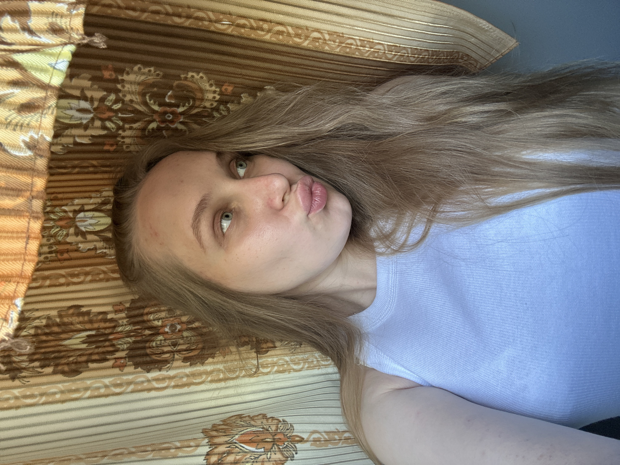 A selfie of a light-skinned young woman with light brown hair and green eyes. She is lying on a patterned brown and beige surface that appears to be a bed or couch. She is wearing a white ribbed tank top. The image provides no information about the city of Chaikovsky, Russia.