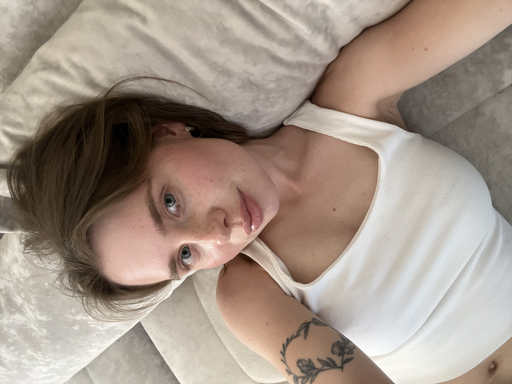 A female individual is depicted in an indoor setting, looking directly towards the viewer. She has medium brown hair, blue eyes, and fair skin with visible freckles across her nose and cheeks. Her lips are full and appear glossy. She is wearing a white ribbed tank top. A tattoo of a floral wreath design is visible on her left upper arm. A small black earring with a silver accent is present in her right ear. She is lying on her back on a light-colored, textured surface, likely a sofa or bed, which features subtle folds. A lighter, smoother pillow or blanket is visible to the right of her head (viewer's left). The specified location for this scene is the city of Chaikovsky, Russia.