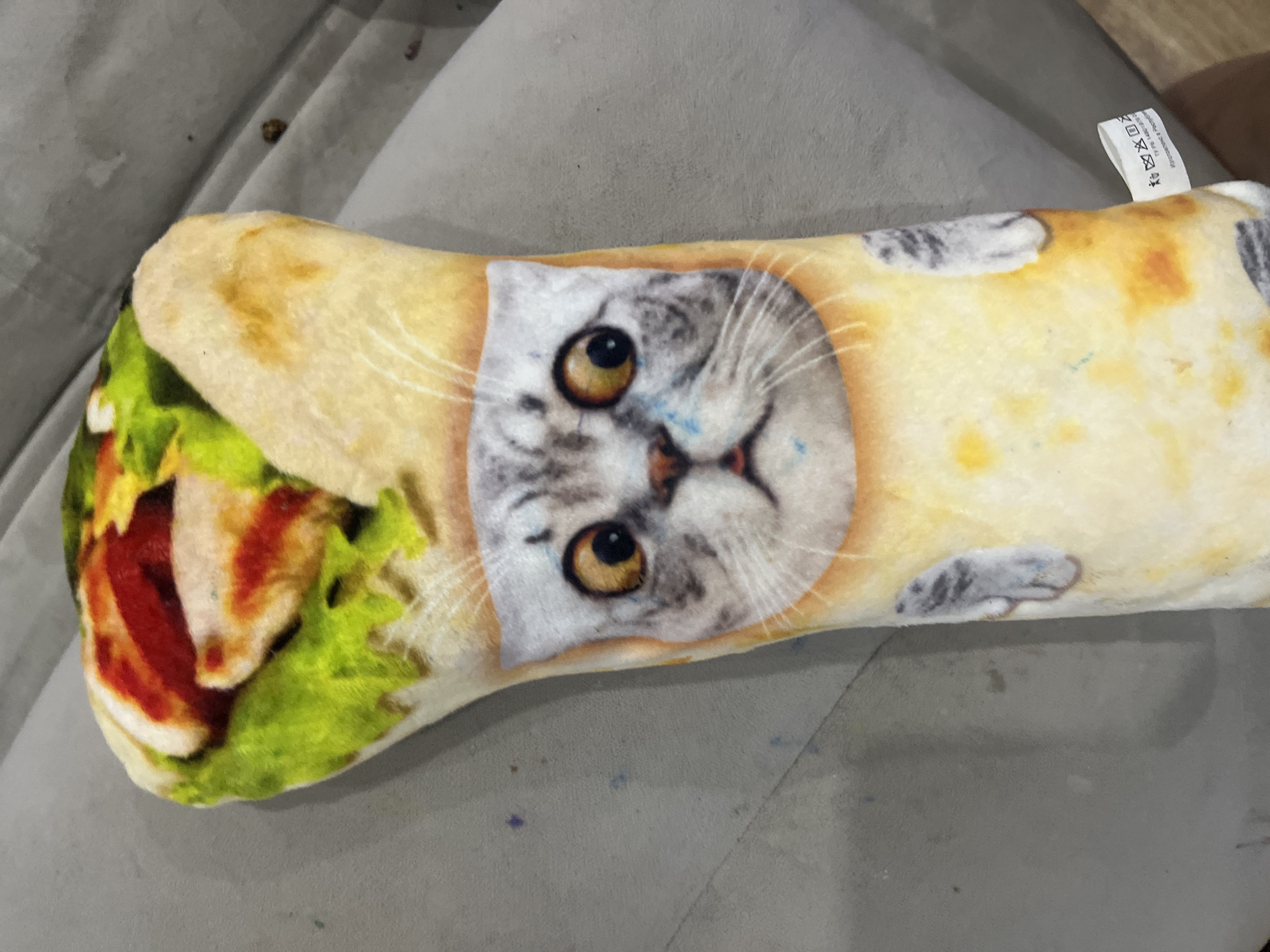 The image shows a stuffed toy shaped like a burrito with a cat's face printed on it. The toy is light-colored, resembling a tortilla, with colorful filling visible at the top, including green lettuce, red sauce, and white meat. The cat's face is centrally located on the toy, featuring large, surprised eyes, gray and white fur markings, and a small pink mouth. Two gray cat paws are depicted on either side of the cat's face. The toy is lying on a textured gray surface, likely upholstery, possibly a couch or chair. A small white tag with laundry symbols and Russian text is attached to the lower right side of the toy. The background is blurred and out of focus, showing a continuation of the gray surface and hints of wood grain on the floor. There is no visible indication of the city of Gorodets, Russia, within the image.