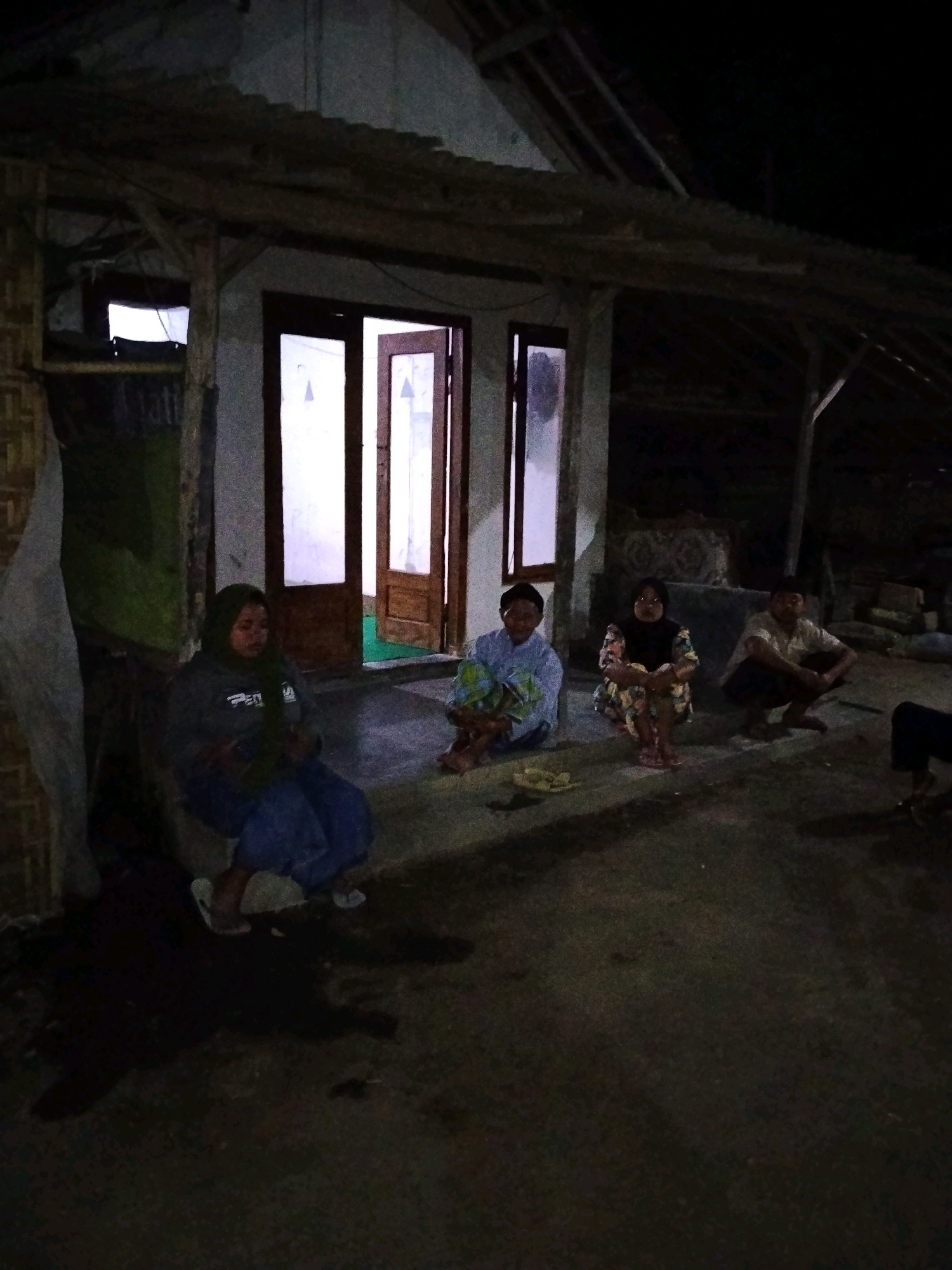 In the city of Tlogosari, Indonesia, four individuals are seated on the ground in front of a rustic, open-air structure at night. The structure features a thatched roof supported by wooden posts and a plastered wall with large, glass-paned doors, two of which are ajar. To the left, a woman wearing a dark hijab and a grey jacket is seated. To her right, a person in a light blue patterned shirt and pants sits with a green bundle on their lap. Next to them, a woman in a colorful floral patterned dress is seated. Further to the right, a man in a light-colored collared shirt and dark pants is crouched. A small plate with what appears to be food is placed in front of the individuals in the center and left of the frame. The ground is a dark, textured surface, possibly concrete or packed dirt, with visible shadows cast from the structure and the people. The overall lighting suggests it is nighttime, with artificial light emanating from within the structure and some ambient illumination.