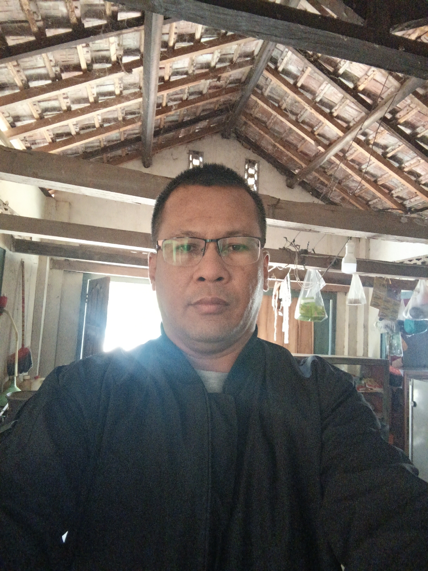 A man wearing glasses and a black jacket is posing for a selfie in what appears to be an indoor, unfinished space. The background shows exposed wooden beams and tiled roofing, indicative of a building under construction or renovation. Sunlight streams in from an open doorway or window to the man's right. Various objects hang from the ceiling and shelves are visible in the background, suggesting a cluttered or utilitarian environment. The man is the sole subject in the frame. There is no explicit indication of the specific location within Gedung Nyapah, Indonesia.