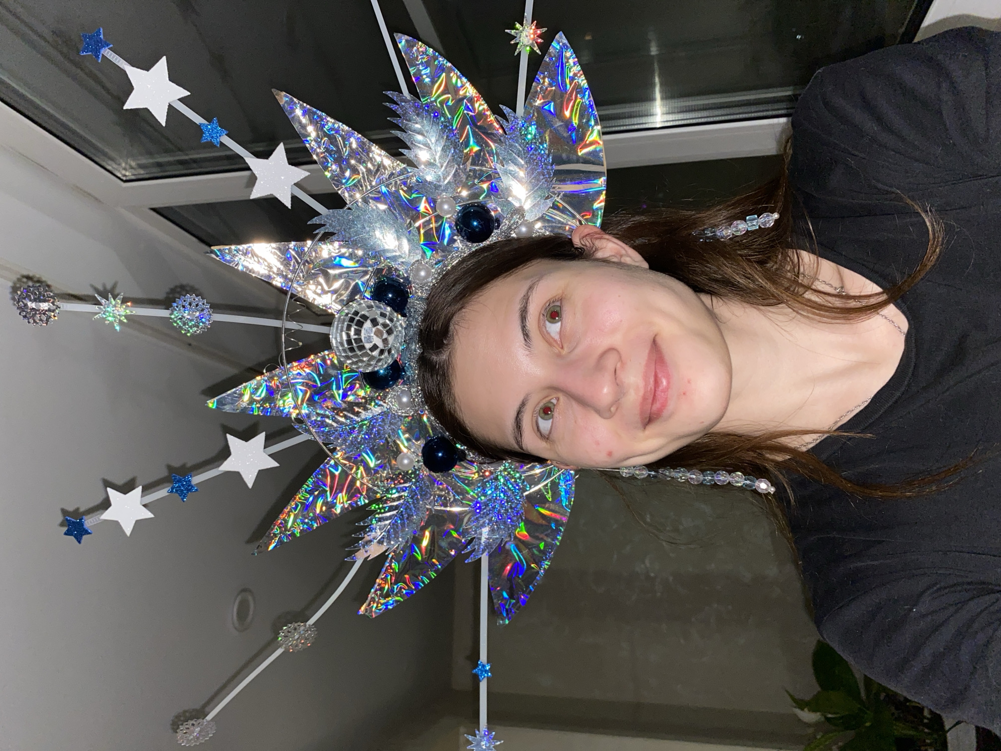 A young woman with brown hair is depicted in a slightly tilted close-up portrait, smiling gently and looking towards the camera. She is wearing a simple black long-sleeved shirt.
Her head is adorned with an elaborate, festive headdress that serves as the central focal point. This intricate piece is constructed from various reflective and glittering materials, predominantly silver and blue. Large, pointed elements made of holographic or iridescent foil reflect a spectrum of rainbow colors. The headdress is further embellished with smaller silver leaf-like pieces, glossy blue and silver spherical beads, and a miniature disco ball. Extending outwards from the main structure are several thin white rods, each tipped with decorative elements such as glittery white stars, smaller blue stars, and intricate snowflake-like ornaments. A string of clear and light blue beads also hangs down, appearing as an elongated earring or hair accessory.
The background suggests an indoor setting, possibly a room, with a window visible on the left side. The walls are a light, neutral color. The lighting appears to be artificial, highlighting the shimmering qualities of the headdress, indicating that the photo was likely taken in the evening or at night. The image conveys a celebratory or creative atmosphere, perhaps for a costume party or event in Votkinsk, Russia. There is no visible text in the image.