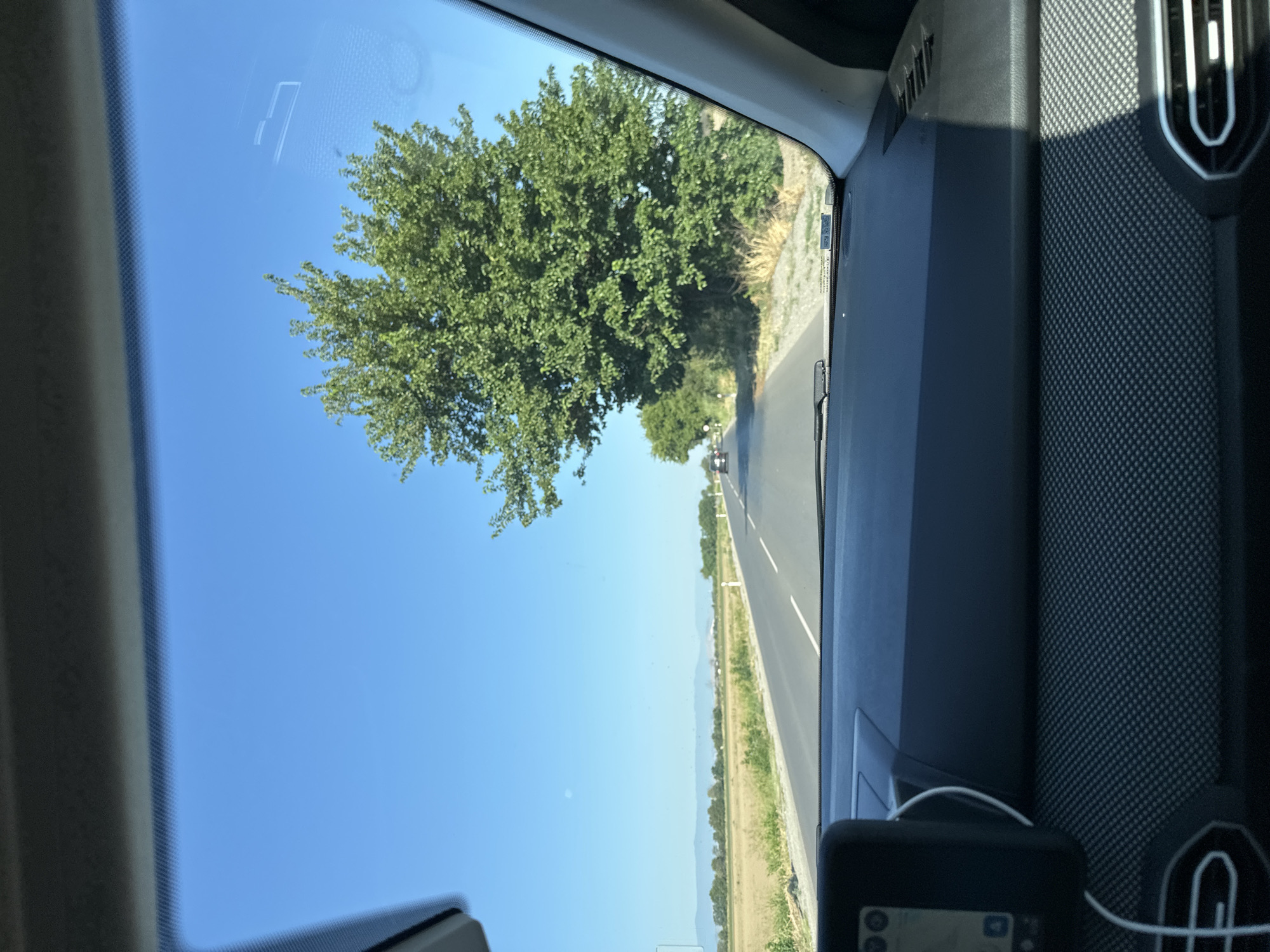 The image is a first-person perspective from inside a car, looking through the windshield. The car is on a paved road with white lane markings, suggesting it is a rural area. The road curves gently to the left and appears to be a two-lane highway.
To the left of the road, there is a field of dry, yellow grass or crops. In the distance, a hazy line of trees and low hills can be seen against a clear, bright blue sky.
To the right of the road, a large, leafy green tree dominates the foreground, its branches extending over the road. Behind this tree, more trees and vegetation line the roadside. The environment appears to be a sunny day in a rural or semi-rural setting.
Inside the car, the dashboard is visible, with a GPS device mounted on the left side. The interior of the car is dark-colored, with a textured surface on the dashboard. The overall impression is a journey through a scenic, open landscape on a clear day.
The location is identified as the city of Orizari, Bulgaria.