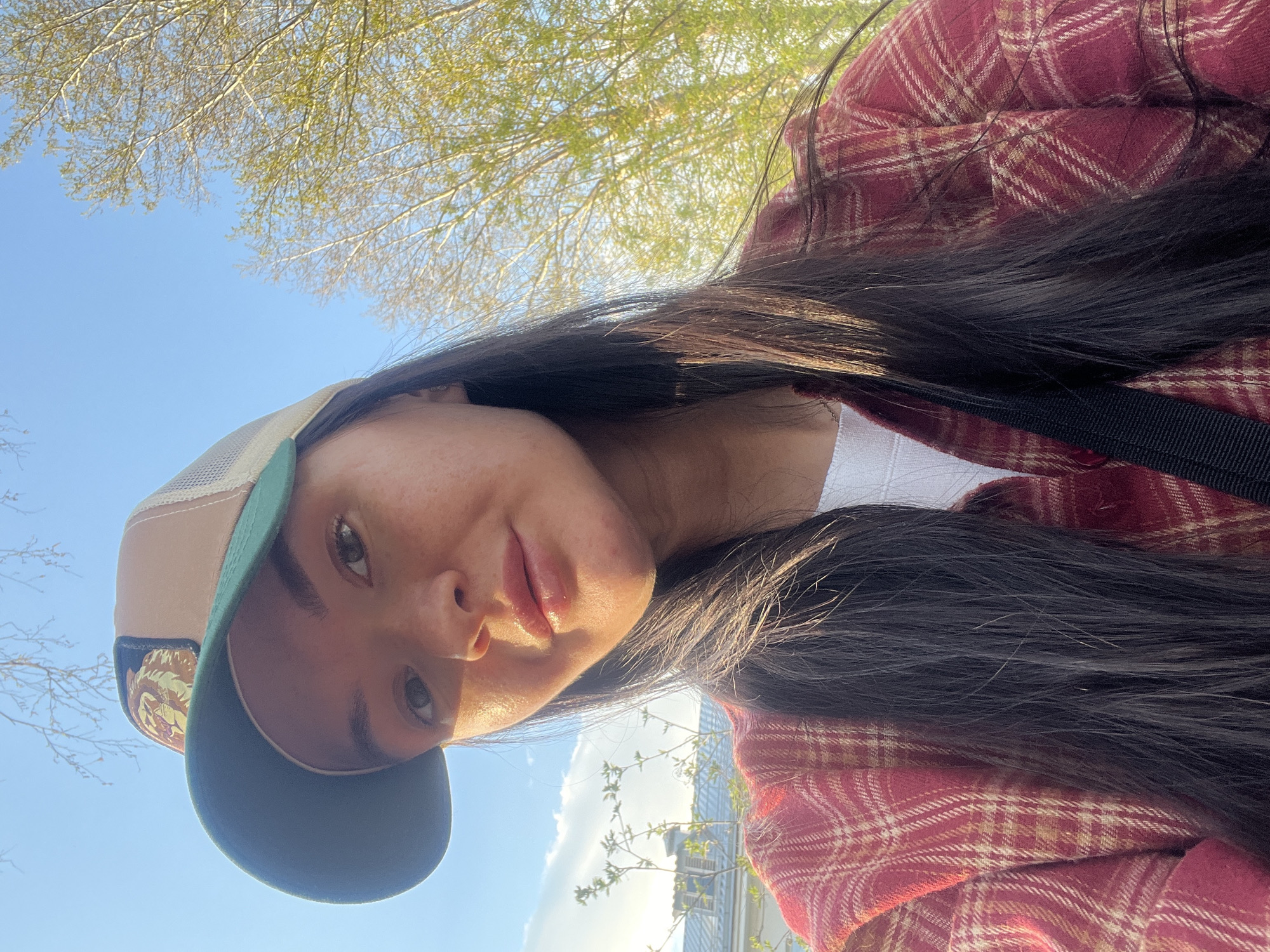 A woman is looking directly at the camera in an outdoor setting. She is wearing a red plaid shirt over a white undershirt, and a cap with a dark green bill and a tan crown. Her long, dark hair is draped over her shirt. The background consists of a clear blue sky partially obscured by budding tree branches. Given the architectural style, the location may be near Kazan, Russia.