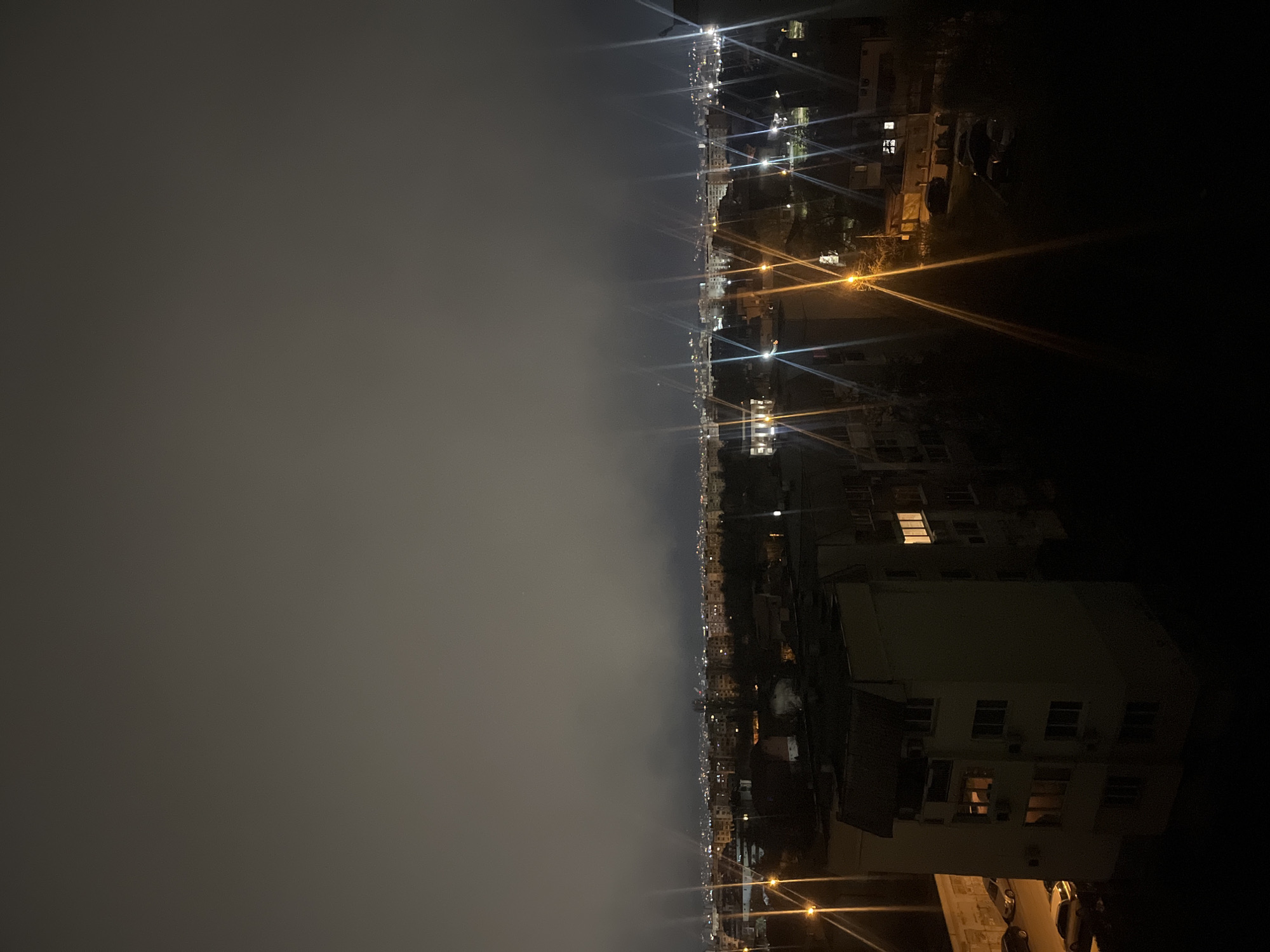 The image shows a nighttime view of Sofia, Bulgaria, from an elevated position, likely a high-rise building. The scene is divided into two contrasting sections: the left side is dominated by a hazy, grey sky, while the right side depicts the cityscape. Buildings are visible, illuminated by streetlights and some interior lights from windows. There are no people visible, but the presence of cars suggests human activity. The general environment is urban, with the city lights stretching into the distance.