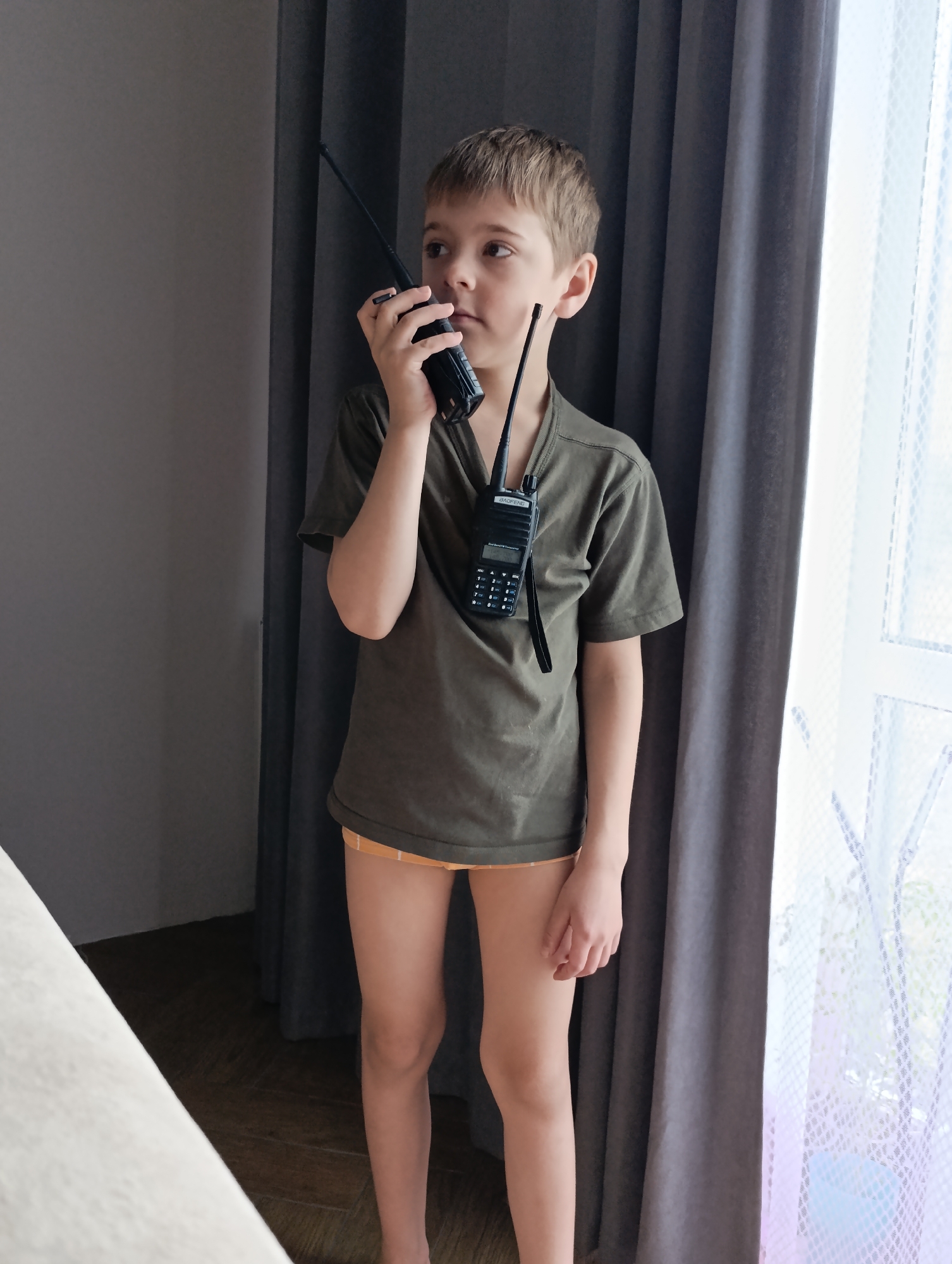 A young boy, appearing to be in elementary school, is depicted indoors, engaged in imaginative play with walkie-talkies. He stands near a window, holding one black walkie-talkie to his mouth as if speaking into it, while another similar walkie-talkie hangs around his neck by a black lanyard. The boy has short, light brown hair and a focused, slightly serious expression, looking towards the right side of the frame.
He is wearing a loose-fitting dark olive green t-shirt and orange and white striped boxer briefs. The walkie-talkie hanging from his neck has the brand "BAOFENG" clearly visible on its display panel, along with a keypad of numbers and functions.
The setting appears to be a residential room. To the boy's left is a plain dark grey wall or curtain. To his right, a large window is visible, covered by sheer white patterned curtains or blinds, with darker grey curtains pulled back on the side. Ample natural light streams in from the window, indicating it is daytime. A partial view of a light-colored surface, possibly a bed or sofa, is visible in the bottom left corner. The floor is dark with a subtle wood pattern. The scene is set in Chaikovsky, Russia.