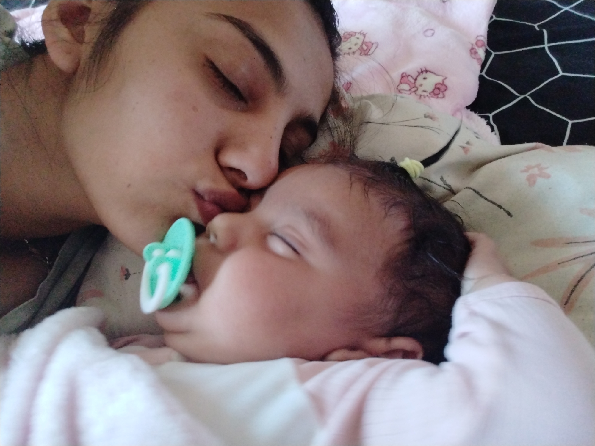 A woman and a baby are lying down indoors. The woman is kissing the baby's head. The baby has a pacifier in its mouth. The location and city are unknown.