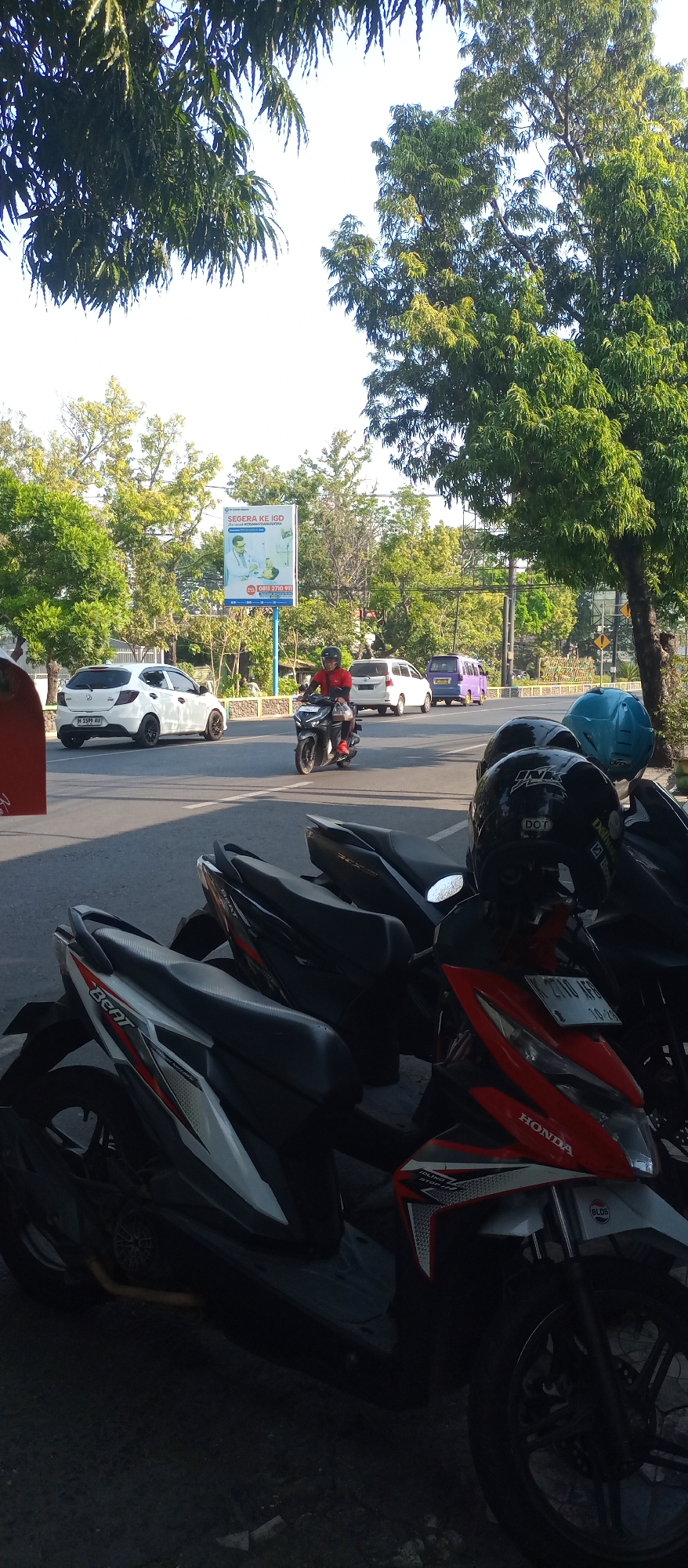 In Jatirejo, Indonesia, on a sunny day, a street scene unfolds. A white Honda Brio car is parked on the left side of a paved road, its license plate reading "M 1399 AV". Further down the road, a man in a red shirt and black helmet rides a black motorcycle. In the background, several other vehicles, including a purple minivan and a white van, are visible. A billboard for a hospital, "SEGERA KE IGD", with an illustration of medical personnel, stands prominently. The foreground is dominated by several parked motorcycles, including a white and red Honda Beat. Two helmets, one black and one blue, rest on the handlebars of one of the motorcycles. Lush green trees line the street, their leaves casting shadows on the pavement. The sky is clear and bright blue.