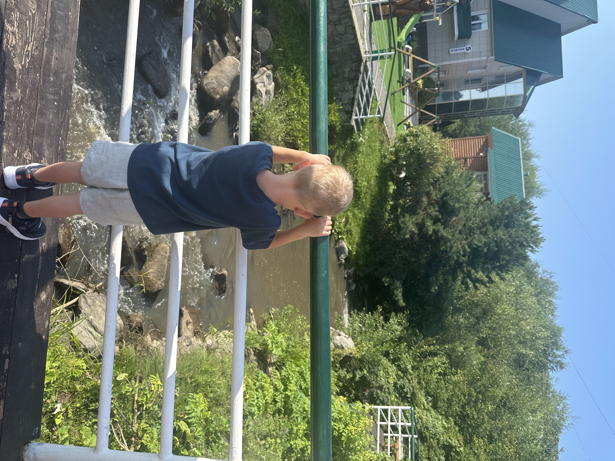 An over-the-shoulder shot shows a young boy with blond hair standing on a wooden bridge in Даниловка, Russia. He is wearing a dark blue t-shirt and light gray shorts. The boy is looking down at a small, rocky river flowing beneath the bridge, with his hands resting on the green railing. The riverbanks are lined with lush green vegetation and rocks. In the background, there are several buildings with green roofs, including a beige, multi-story structure with a glass-enclosed section and a smaller, brown wooden building. A playground with swings and a slide is visible on the left side of the frame. The sky is clear and blue.