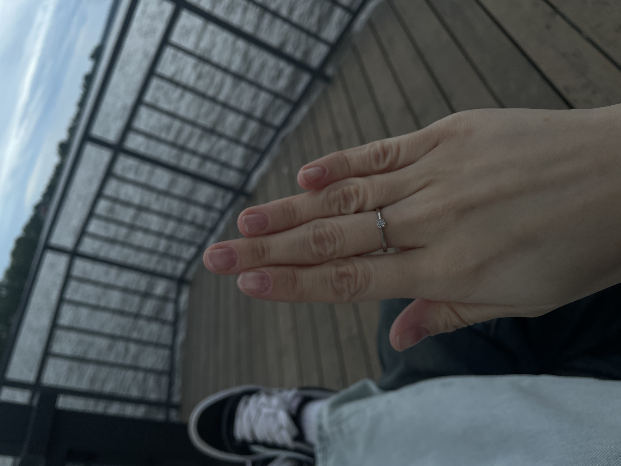 An outdoor scene depicts a person's left hand and lower leg against a waterfront backdrop, assumed to be in Votkinsk, Russia. The hand, with natural-toned nails, is held up to the camera, prominently displaying a slender silver-colored ring featuring a single small, clear round stone on the ring finger. To the left, a portion of the person's leg is visible, clad in light-colored jeans, ending in a black and white sneaker with white laces. The individual appears to be seated on a wooden deck, characterized by parallel planks. A dark metal railing with vertical bars separates the deck from a calm body of water, likely a lake or river. The background consists of the water under an overcast sky, with a faint, distant tree line indicating a far shore.